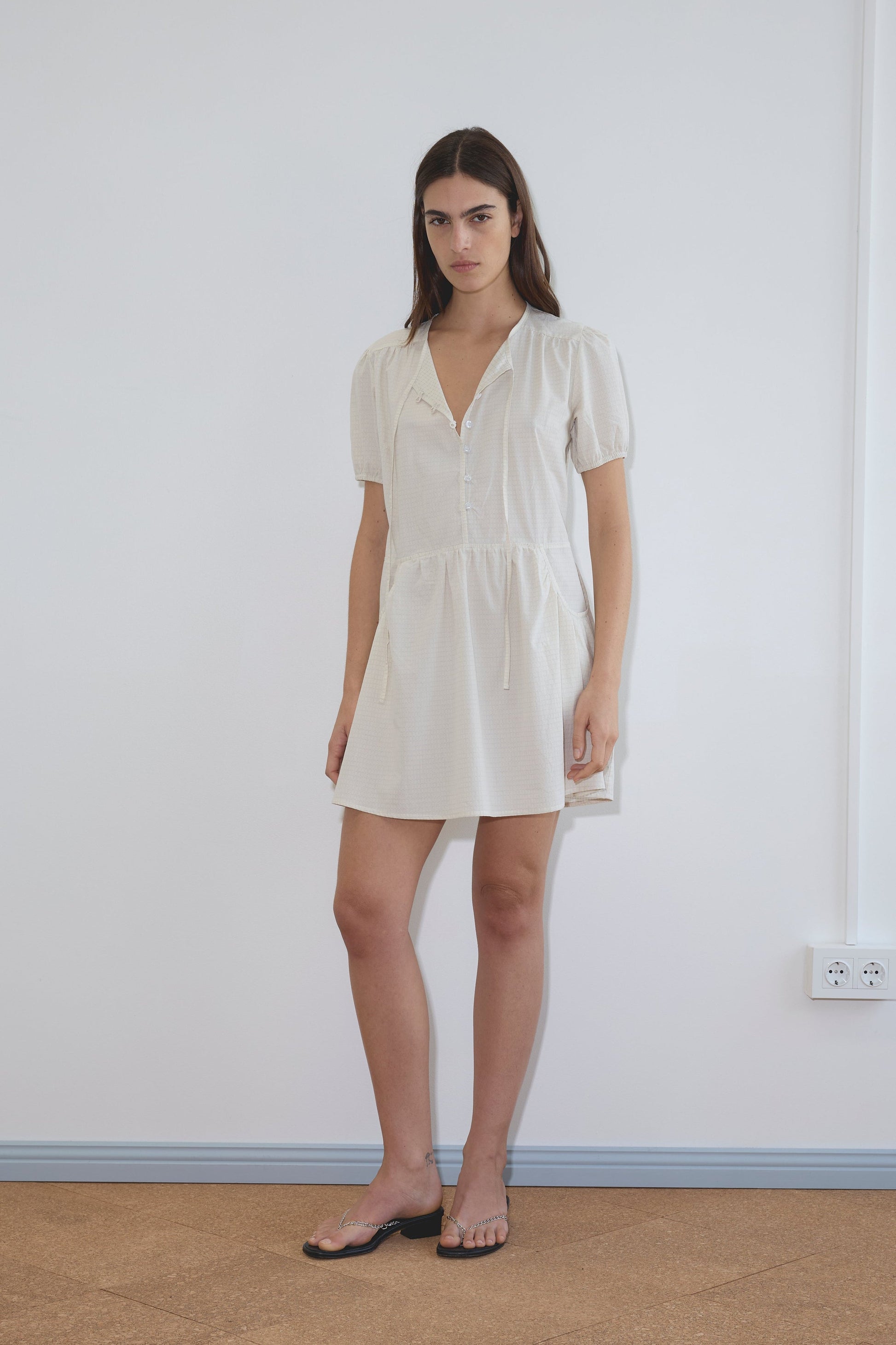 A woman stands indoors against a plain white wall, wearing the Pocket Mini Dress - Bone Check with short sleeves and black sandals. She has long, dark hair and a neutral expression.
