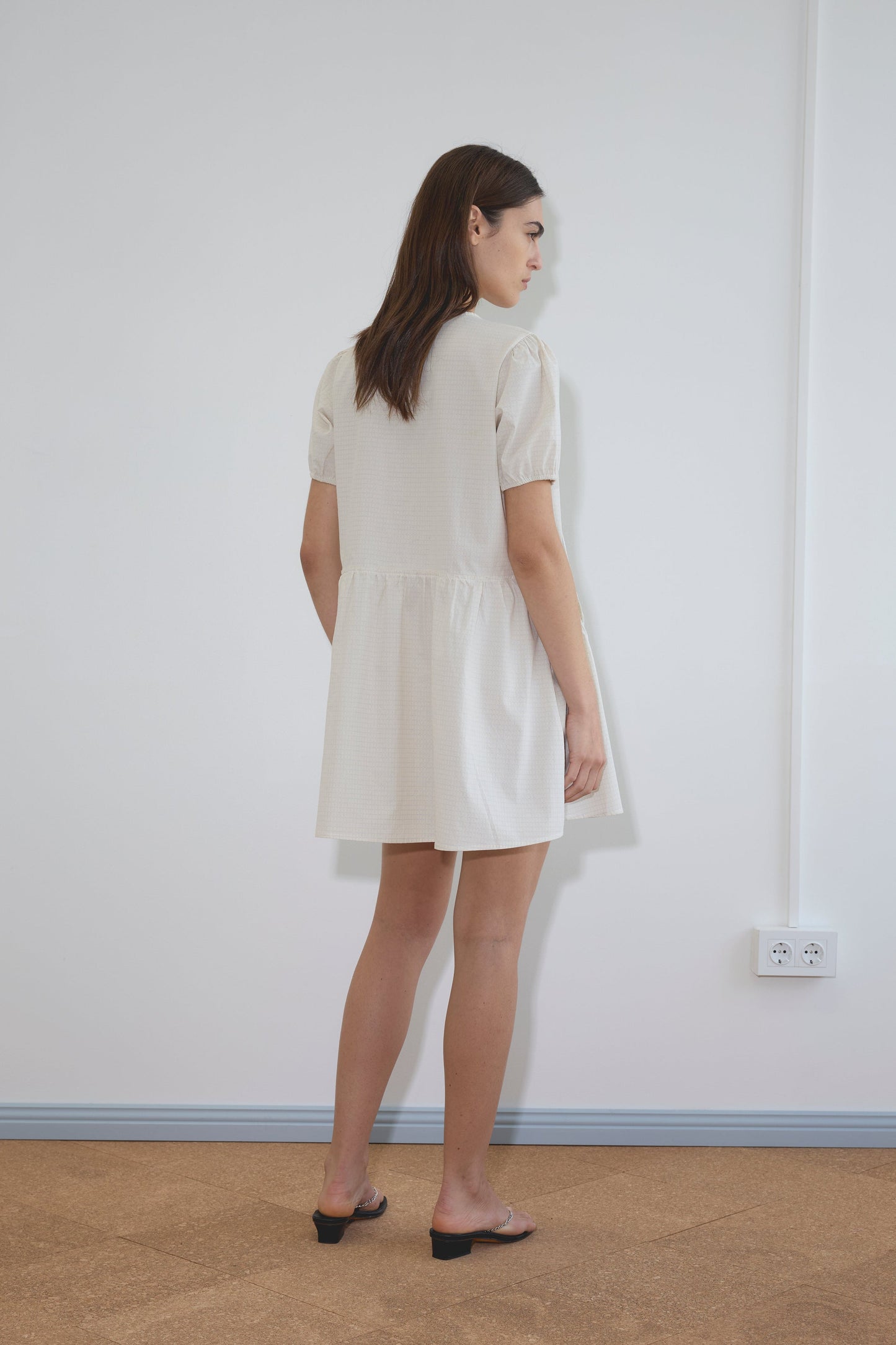 A woman with long brown hair stands indoors facing a wall, wearing the Deiji Studios Pocket Mini Dress - Bone Check with short sleeves and black heeled sandals. A white electrical outlet is visible on the wall.
