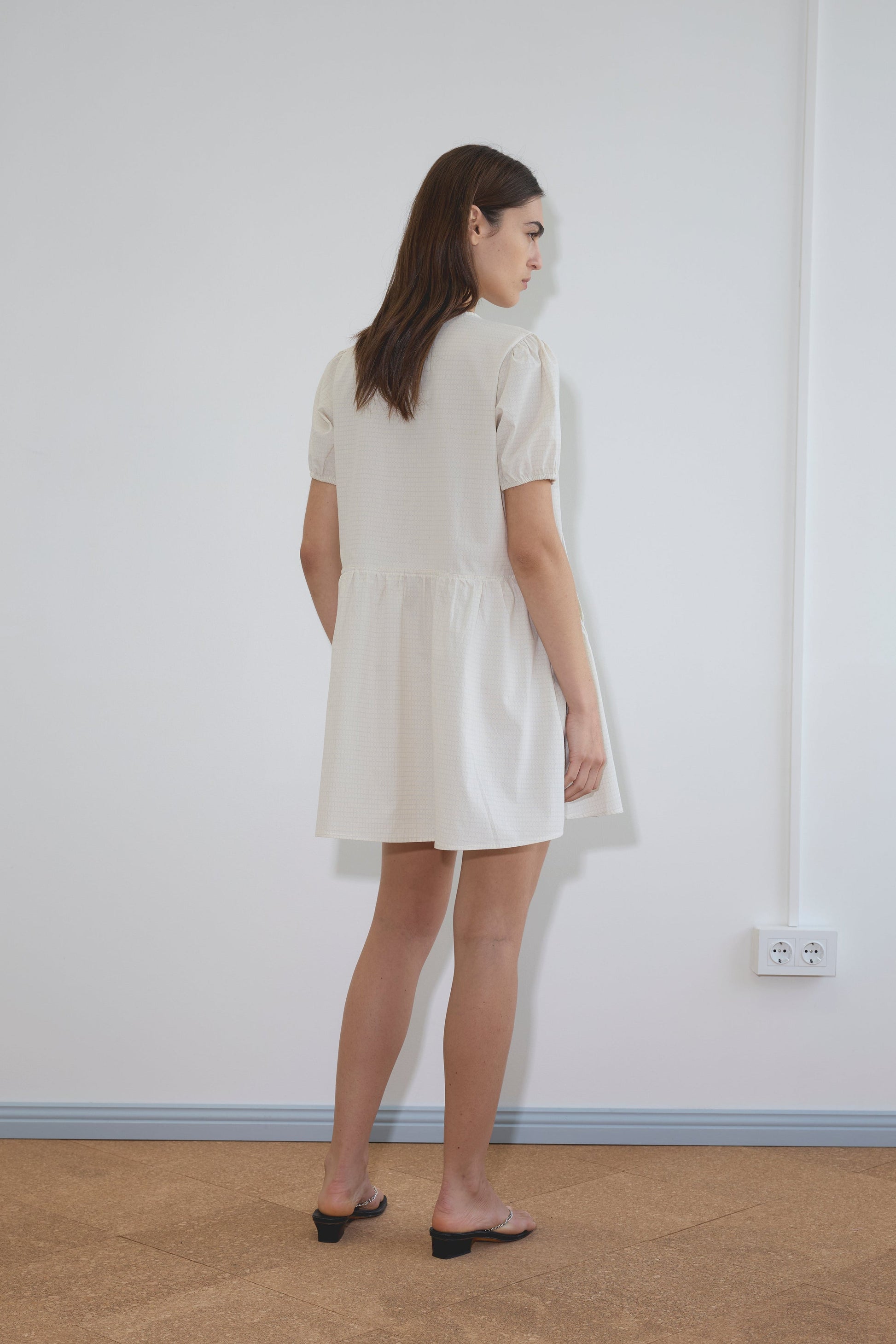 A woman with long brown hair stands indoors facing a wall, wearing the Deiji Studios Pocket Mini Dress - Bone Check with short sleeves and black heeled sandals. A white electrical outlet is visible on the wall.