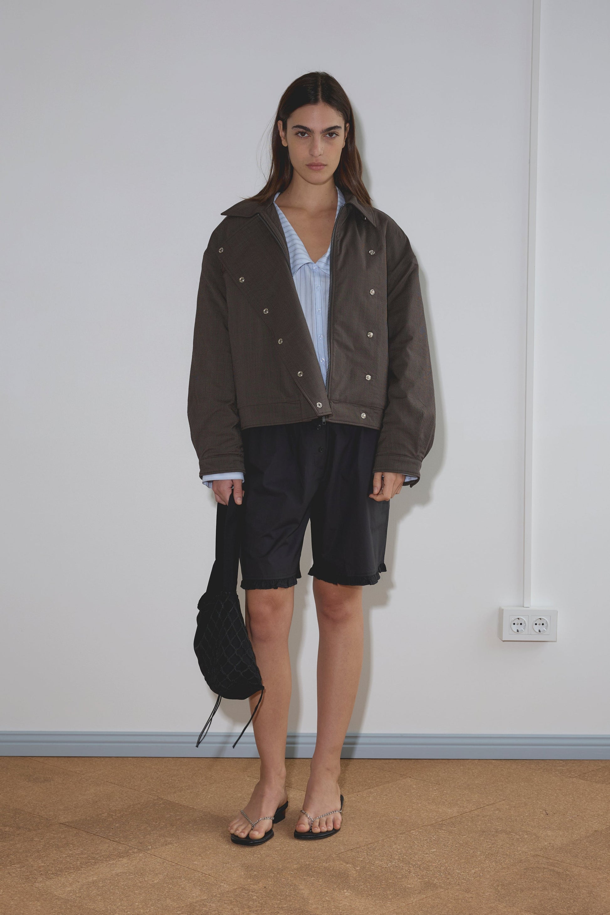 A person with long dark hair stands indoors in a Lapel Jacket - Brown Stripe over a blue shirt, black shorts, and flip-flops, holding a black bag. The plain white background includes a visible electrical outlet.
