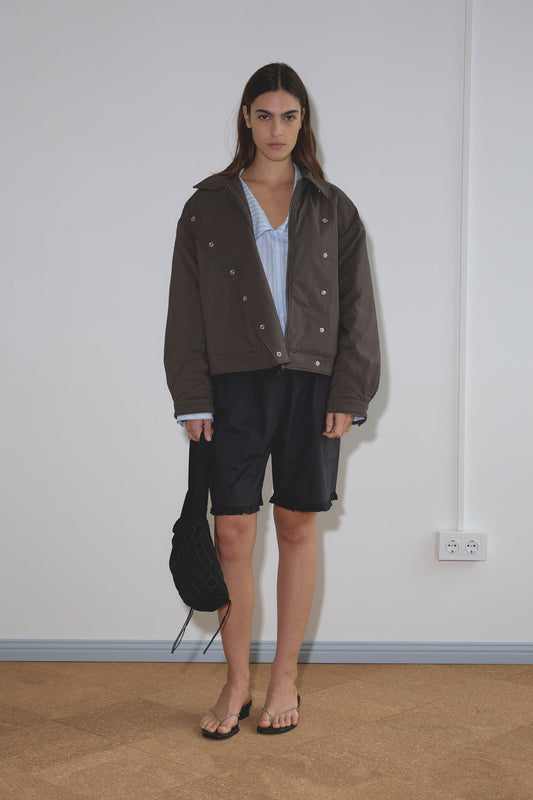 A person with long dark hair stands indoors in a Lapel Jacket - Brown Stripe over a blue shirt, black shorts, and flip-flops, holding a black bag. The plain white background includes a visible electrical outlet.