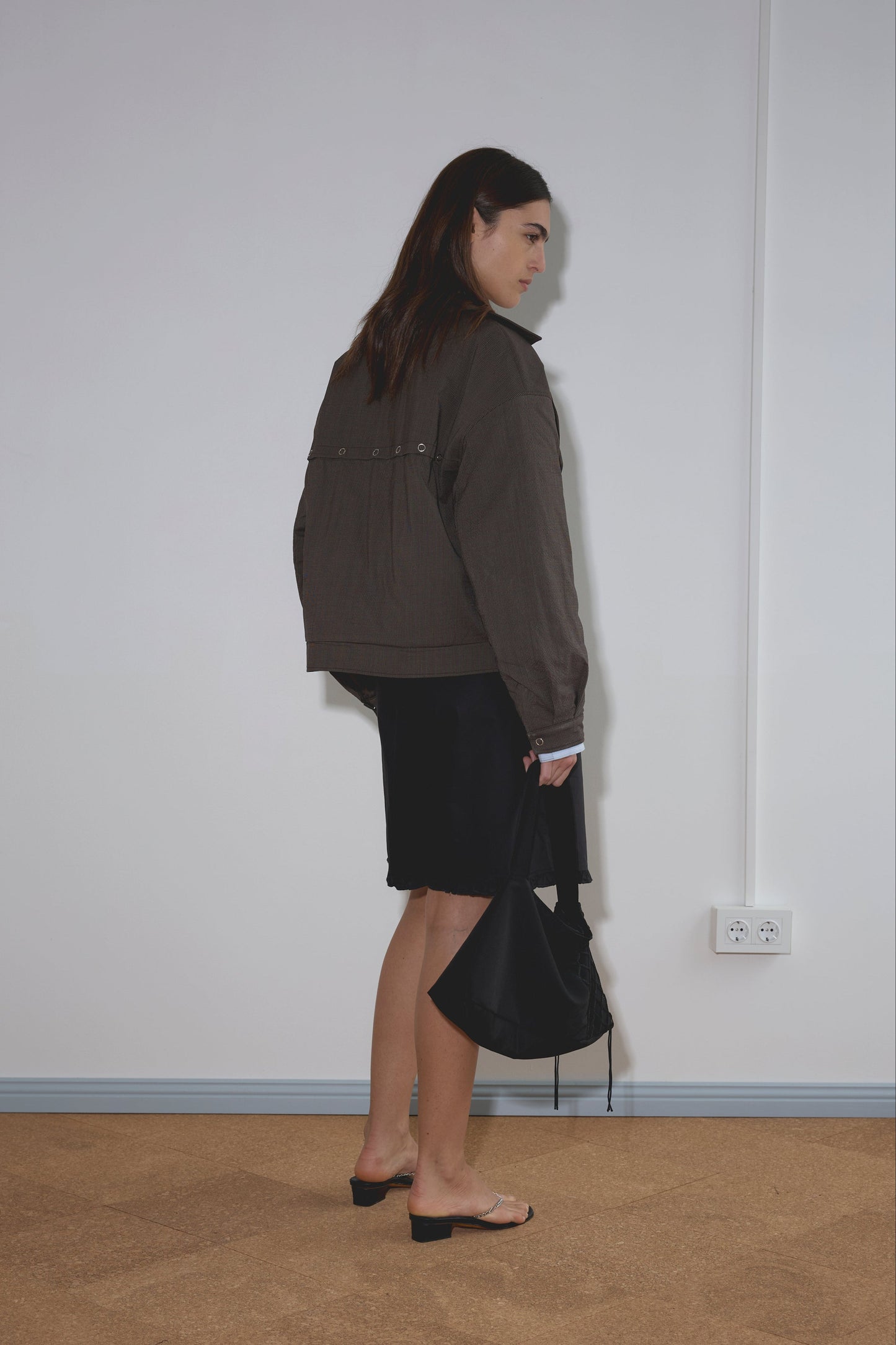 A woman with long brown hair stands sideways indoors, wearing the Lapel Jacket - Brown Stripe from Deiji Studios, a black skirt, and heeled sandals, holding a black handbag and facing a plain white wall with floor outlets.