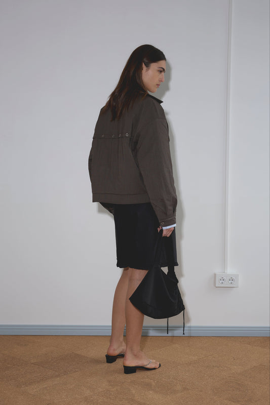 A woman with long brown hair stands sideways indoors, wearing the Lapel Jacket - Brown Stripe from Deiji Studios, a black skirt, and heeled sandals, holding a black handbag and facing a plain white wall with floor outlets.