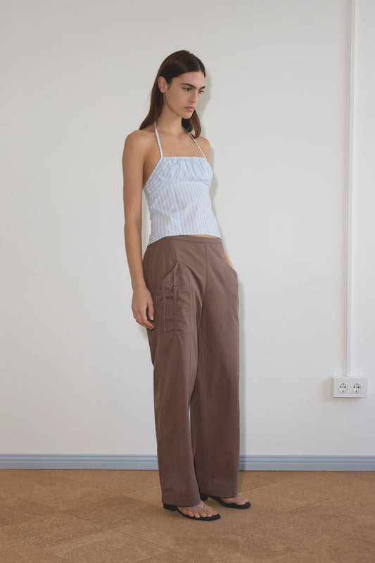 A woman stands indoors, hands in pockets, wearing the Strap Top - Mineral Stripe and loose brown cargo pants. She looks slightly downward against a plain white wall with visible electrical outlets.