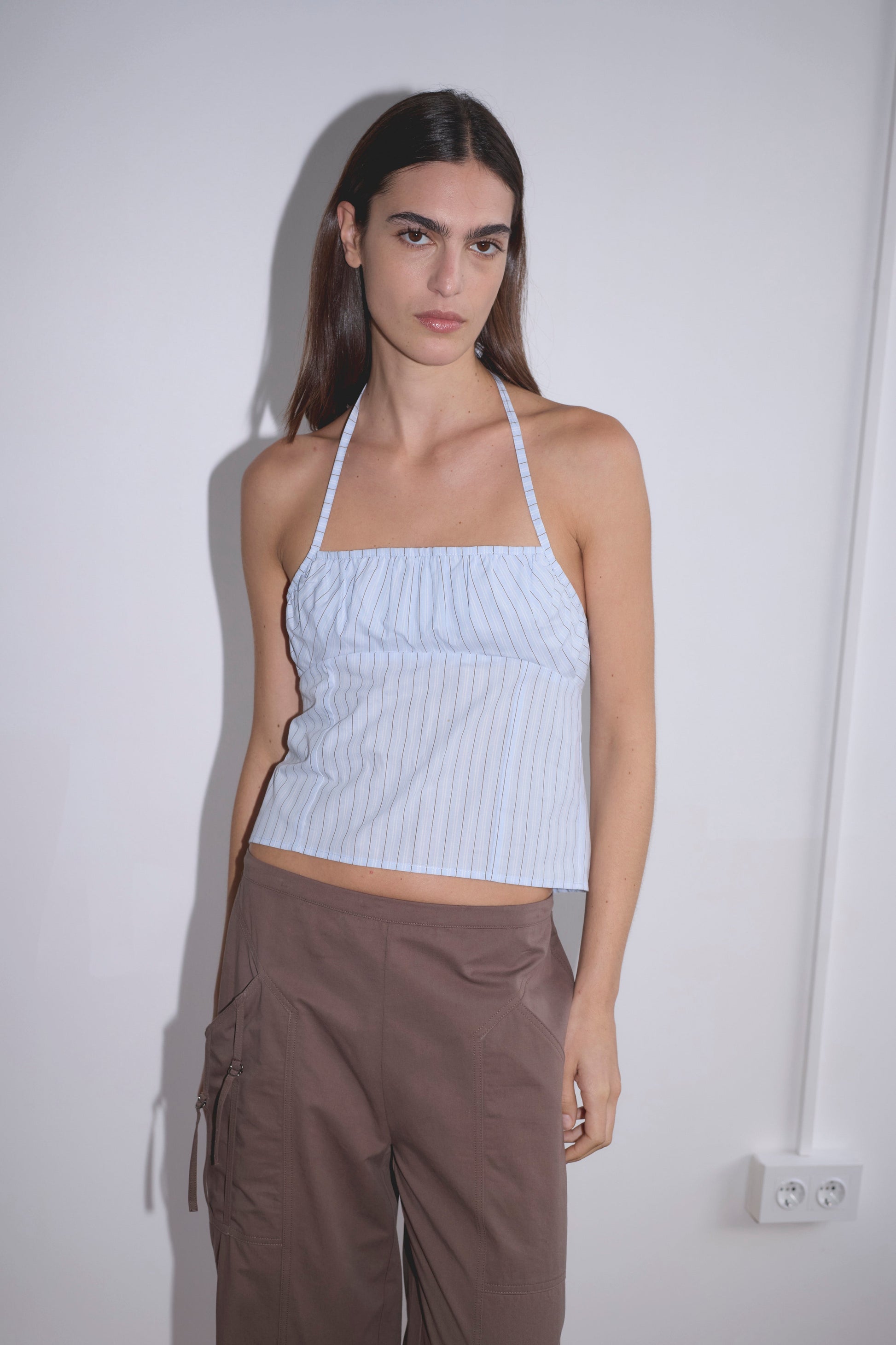 A person with long dark hair stands against a plain white wall wearing the Strap Top - Mineral Stripe, a light blue striped organic cotton halter top, paired with brown cargo pants. An electrical outlet is visible on the wall.