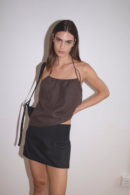 A woman with straight brown hair wears the O Ring Halter - Bark and a black mini skirt, standing against a plain white wall with one hand on her hip, looking at the camera.