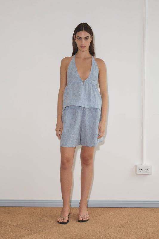 A woman stands indoors against a white wall, wearing the Linen Button Halter - Blue Stripe by Deiji Studios with matching loose shorts in soft French linen. She has long dark hair and is also wearing black sandals.