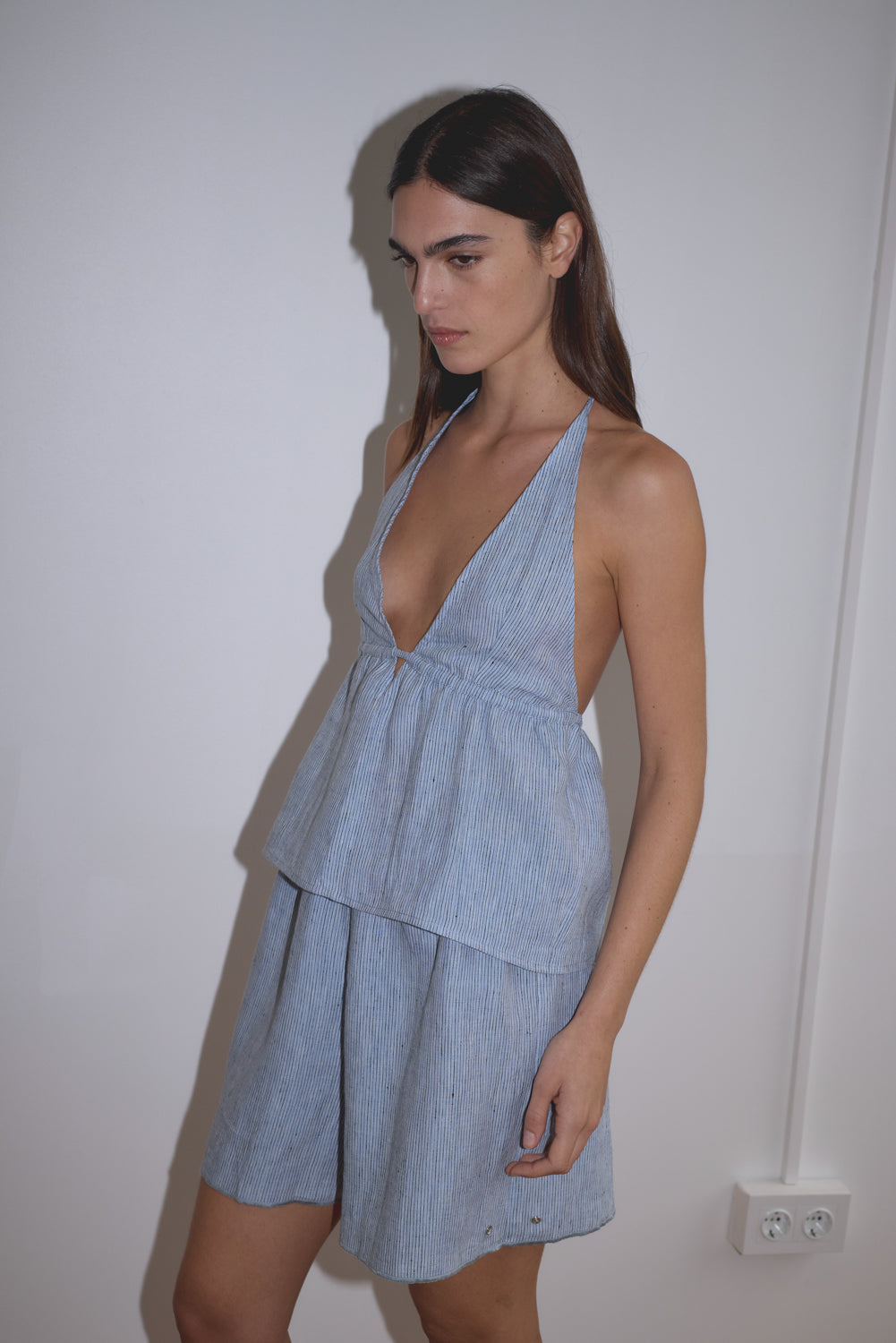 A woman with long dark hair stands against a plain white wall, wearing the Press Stud Short in blue stripe, a light blue, sleeveless piece featuring a V-neck and layered skirt, crafted from soft French linen.