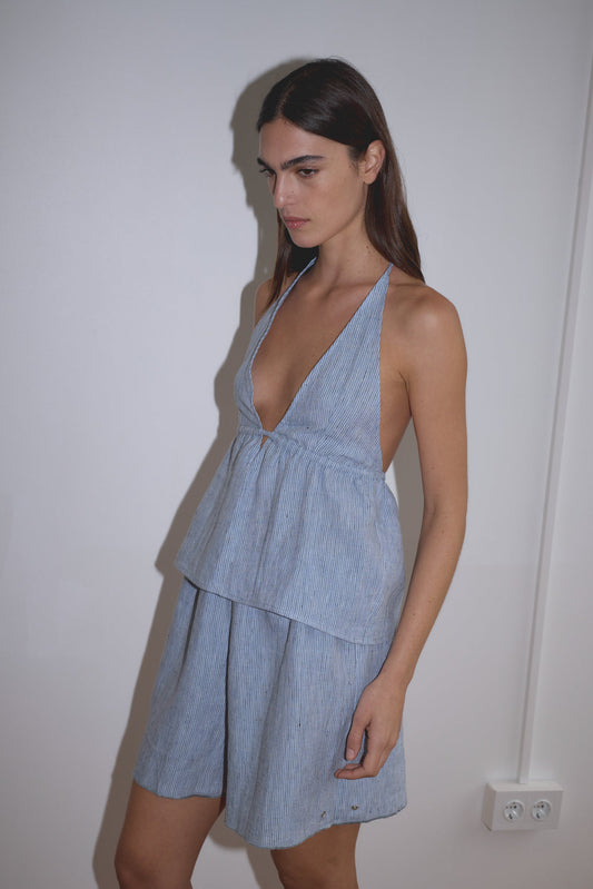 A woman with long dark hair stands against a plain white wall, wearing the Press Stud Short in blue stripe, a light blue, sleeveless piece featuring a V-neck and layered skirt, crafted from soft French linen.