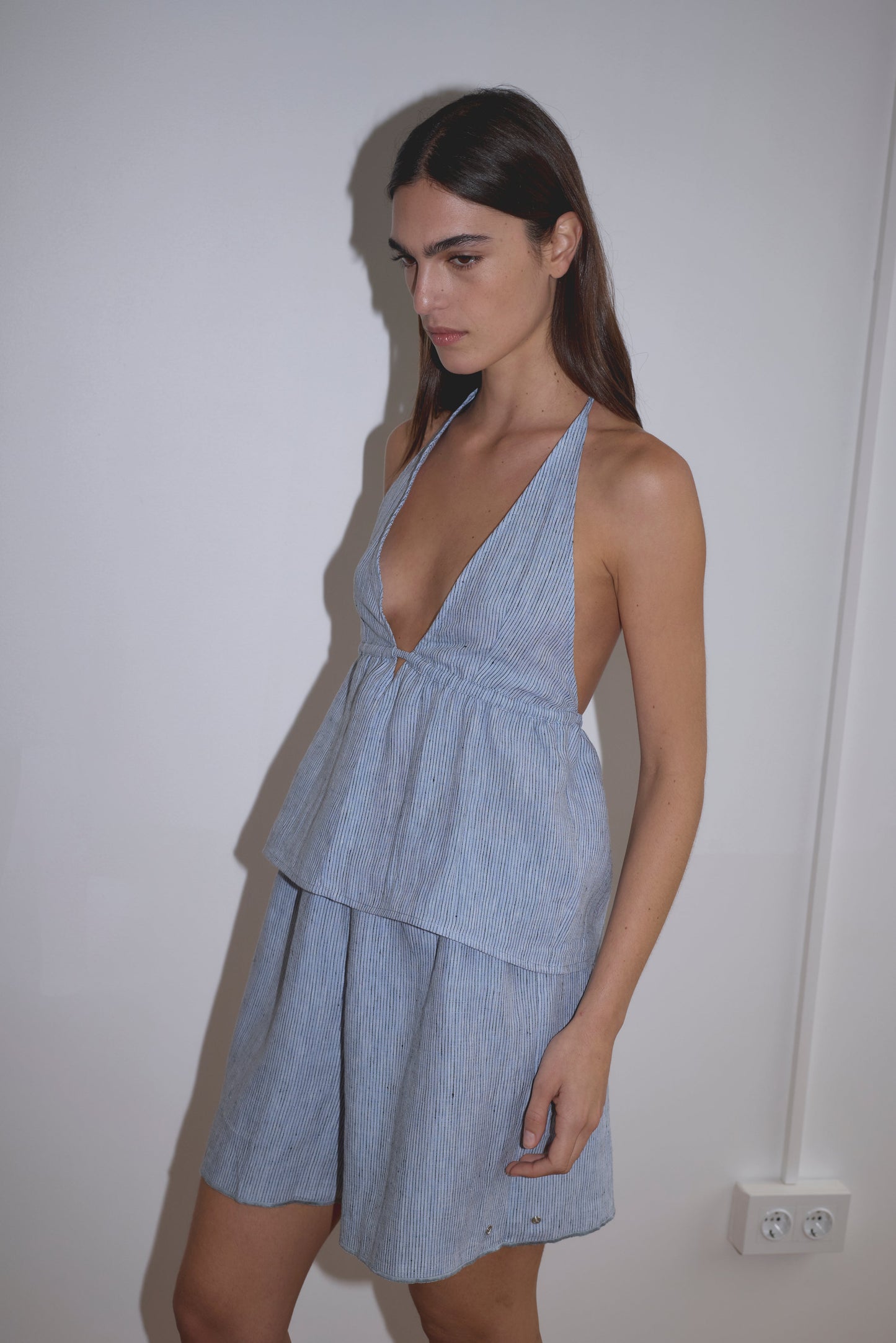 A person with long dark hair wears the Linen Button Halter - Blue Stripe by Deiji Studios, a sleeveless dress with a deep V-neck and flared skirt, standing against a plain white wall.