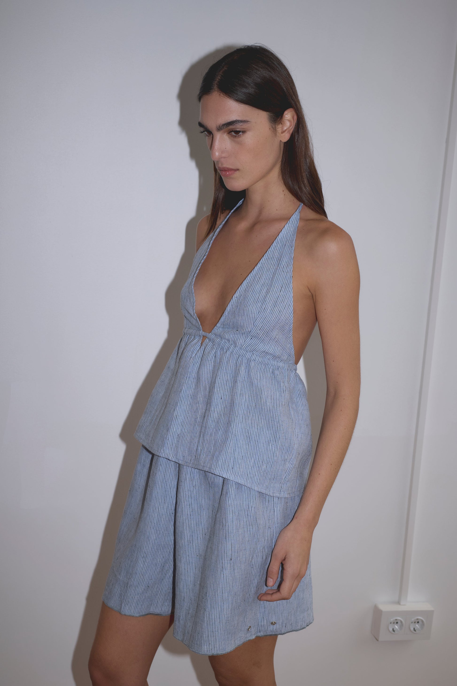 A person with long dark hair wears the Linen Button Halter - Blue Stripe by Deiji Studios, a sleeveless dress with a deep V-neck and flared skirt, standing against a plain white wall.