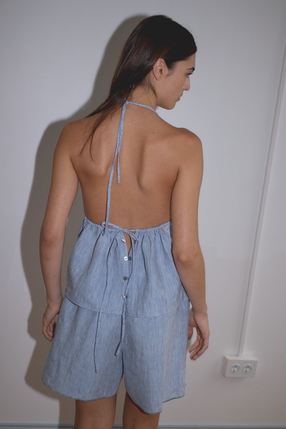 A woman with loose brown hair stands near a light-colored wall, wearing Deiji Studios' Press Stud Short in Blue Stripe—a backless halter dress with buttons down the back, crafted from soft French linen.