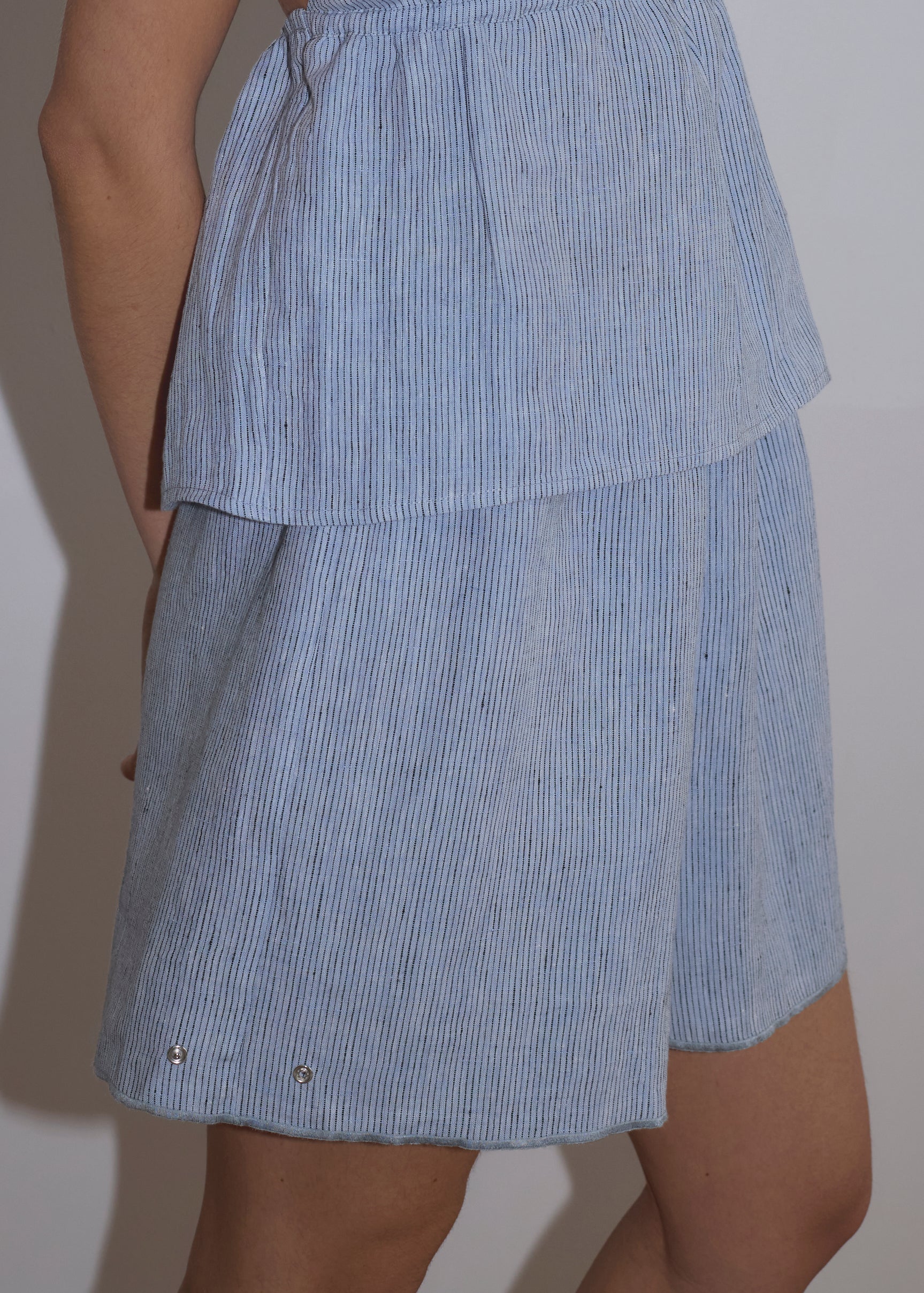 A person wearing Press Stud Shorts with a light blue and white vertical stripe pattern, paired with a matching sleeveless top, stands against a plain light background, showcasing a relaxed, summery style.