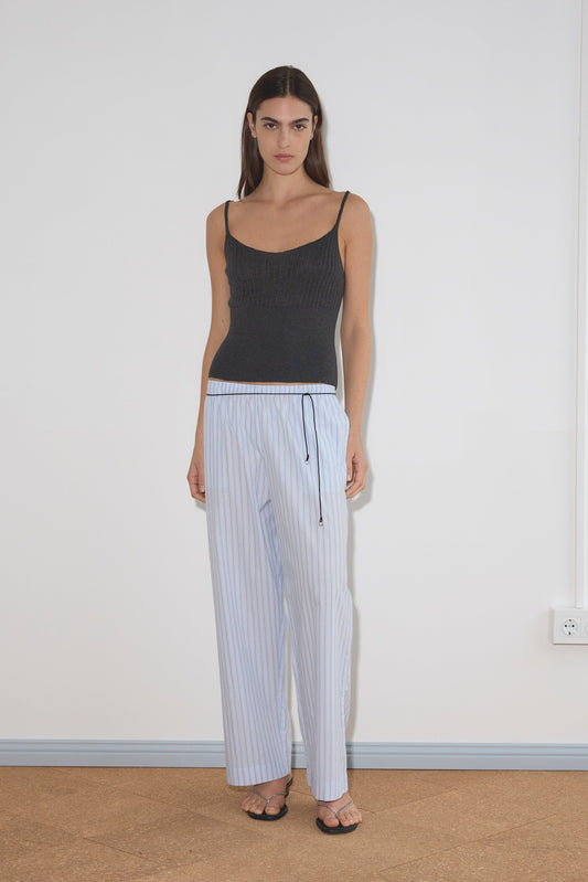 A model wears a dark grey ribbed camisole, Deiji Studios' Ease Trouser - Mineral Stripe (light blue pinstripe, ankle length), and black chain-strap sandals, standing against a white wall.