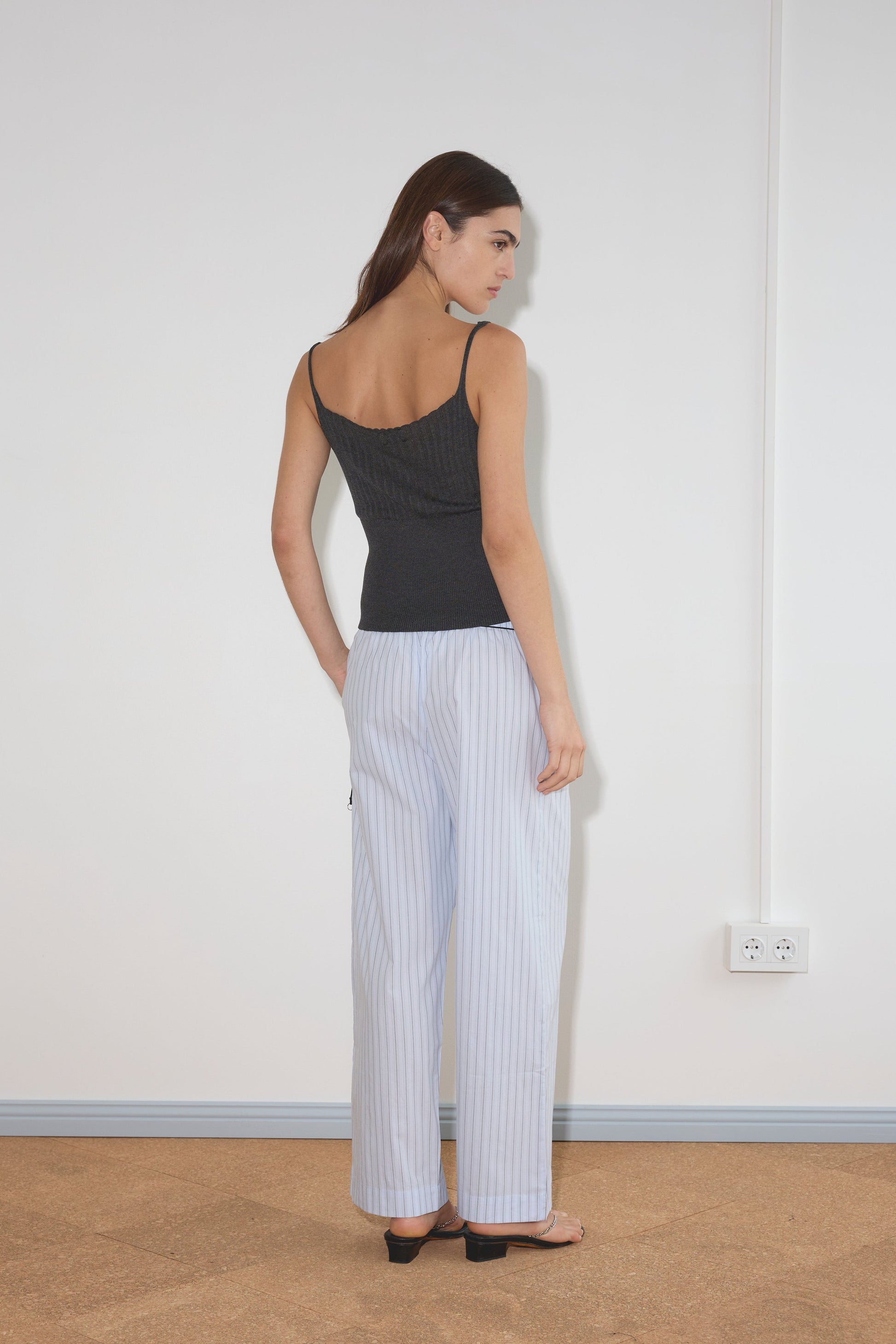 A woman faces away, wearing a black sleeveless top and Deiji Studios Ease Trouser - Mineral Stripe in organic cotton with black sandals, standing against a plain white wall with a visible power outlet.