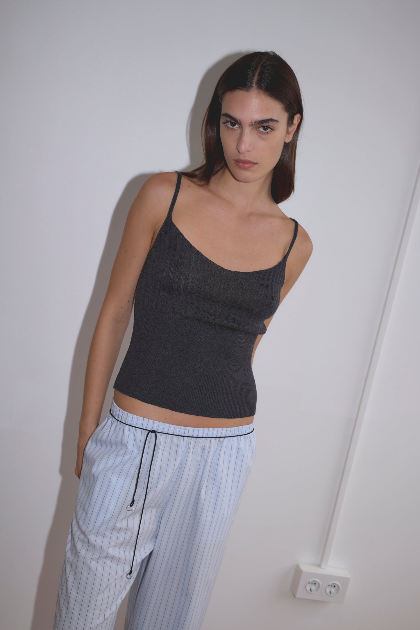 A person with straight, dark hair wears a Deiji Studios Ribbed Tank - Dark Charcoal and light blue striped pajama pants, standing against a white wall with a power outlet visible at the bottom.