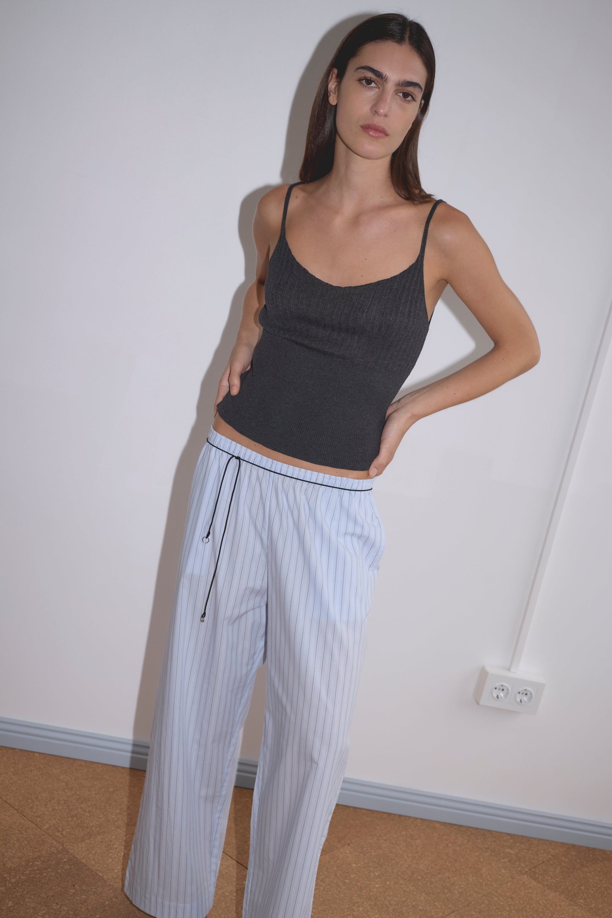 A woman with long, straight brown hair and a neutral expression stands against a white wall, wearing a dark gray sleeveless top and Deiji Studios’ Ease Trouser - Mineral Stripe in loose, light blue pinstriped organic cotton.