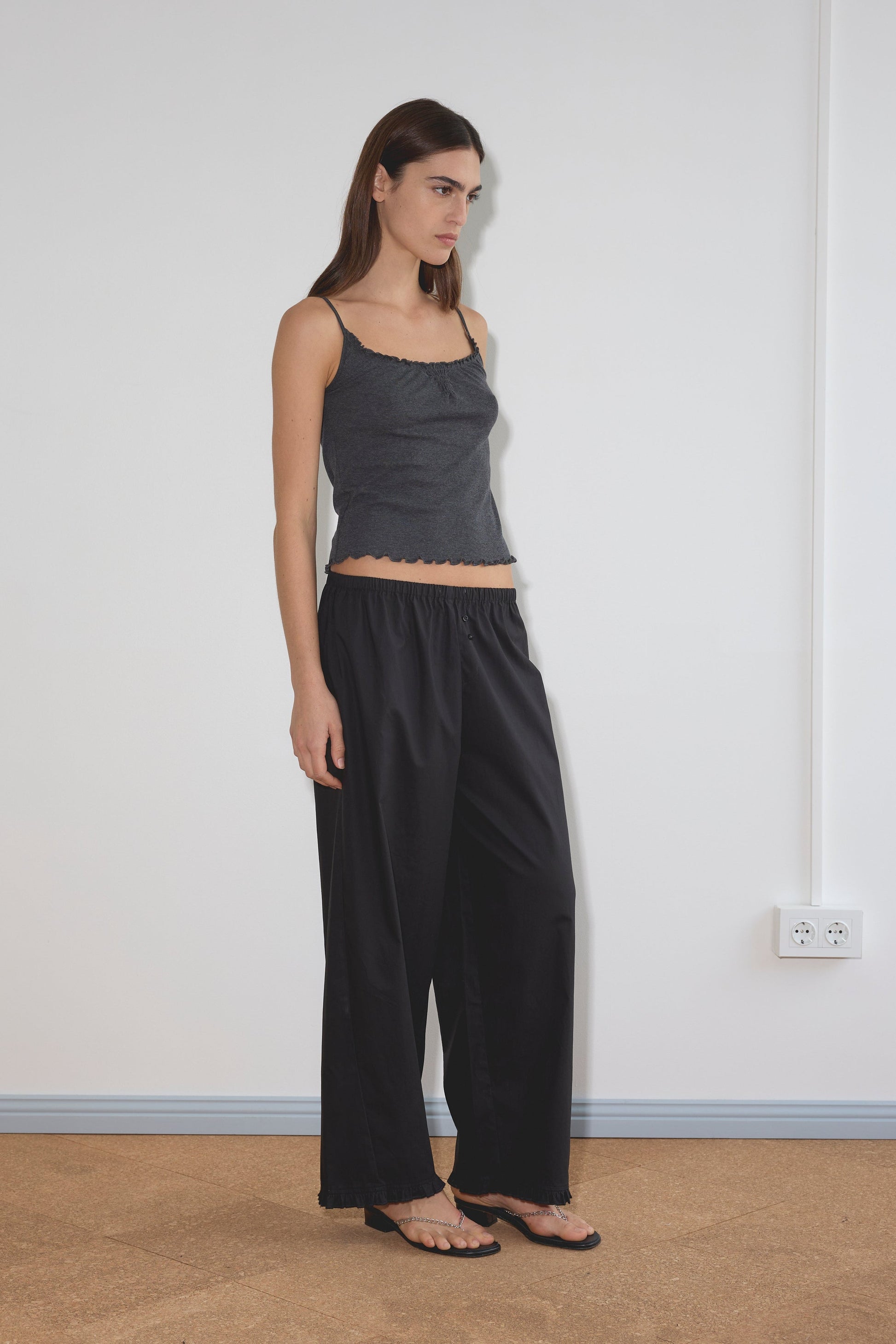 A woman stands indoors against a white wall, wearing the Flounce Pant - Black. She has on a dark gray sleeveless top and black sandals, with straight brown hair and a neutral expression, her arms relaxed by her sides.
