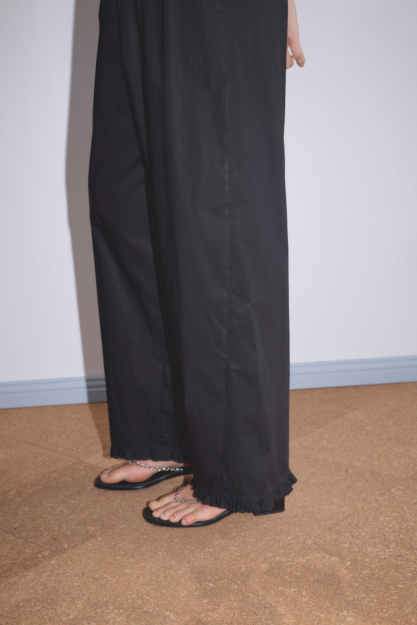 A person wearing Deiji Studios' Flounce Pant - Black and black open-toe sandals stands on a brown cork floor near a white wall with a light blue baseboard. Only their lower legs and feet are visible.