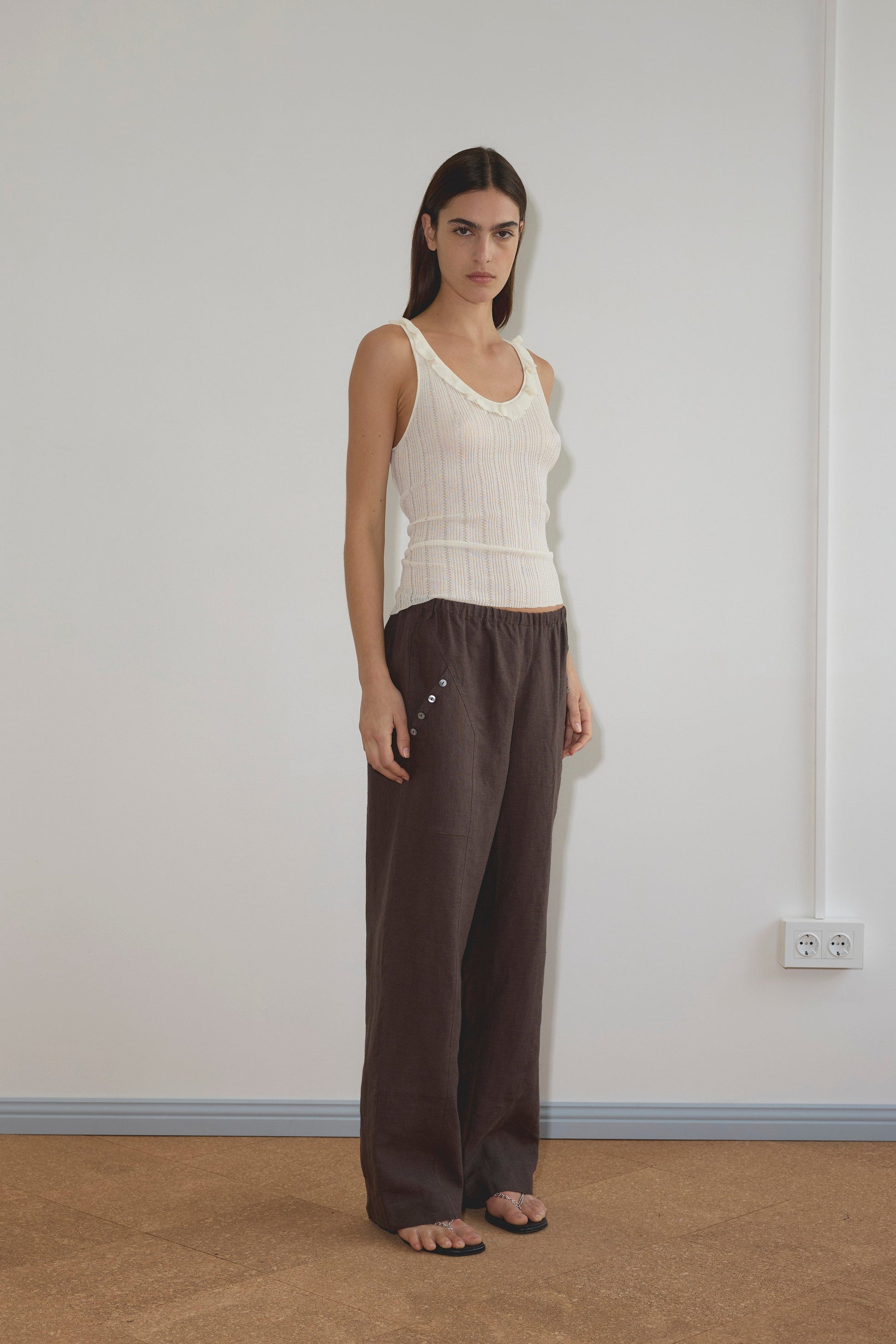 A woman with straight dark hair wears a sleeveless white knit top and the Angle Pocket Pant - Bark, a relaxed mid-waist pant with side buttons, standing barefoot on a brown floor against a plain white wall.
