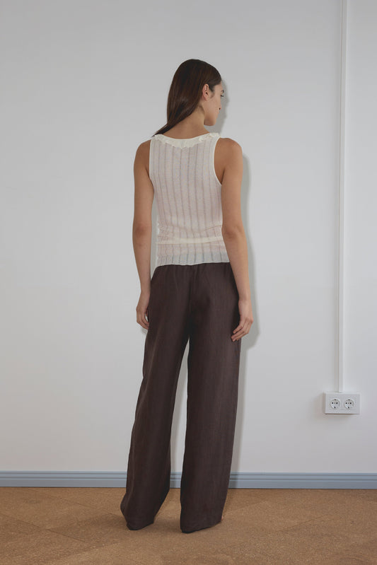 A woman with long brown hair stands facing a white wall, wearing a sleeveless white knit top and the Angle Pocket Pant in Bark. A white power outlet is visible on the wall to her right.
