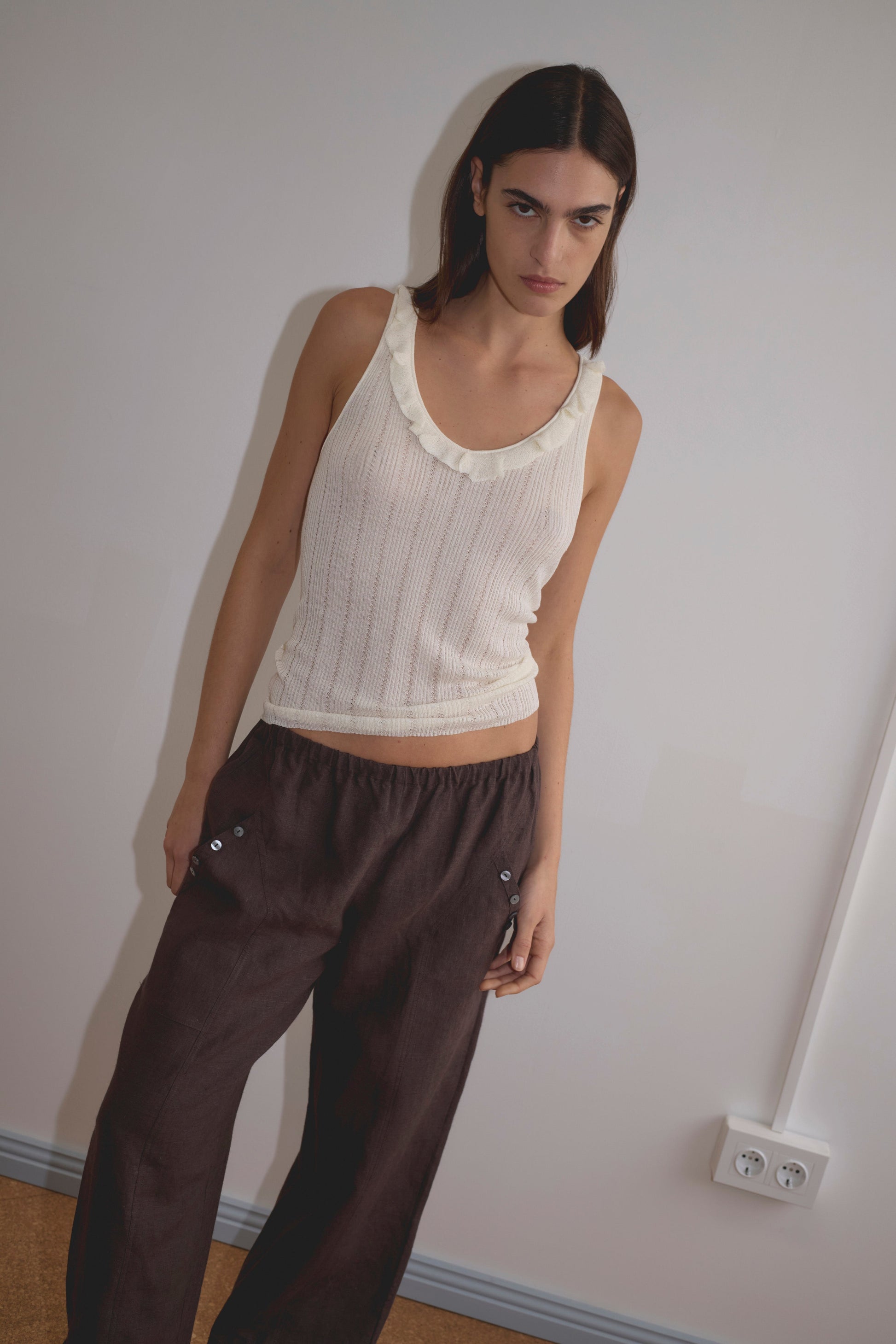 A woman with long brown hair stands against a white wall, wearing a sleeveless cream top and the Angle Pocket Pant - Bark in organic linen with a relaxed mid-waist fit and angled pocket. She has a neutral expression, one hand in her pocket.