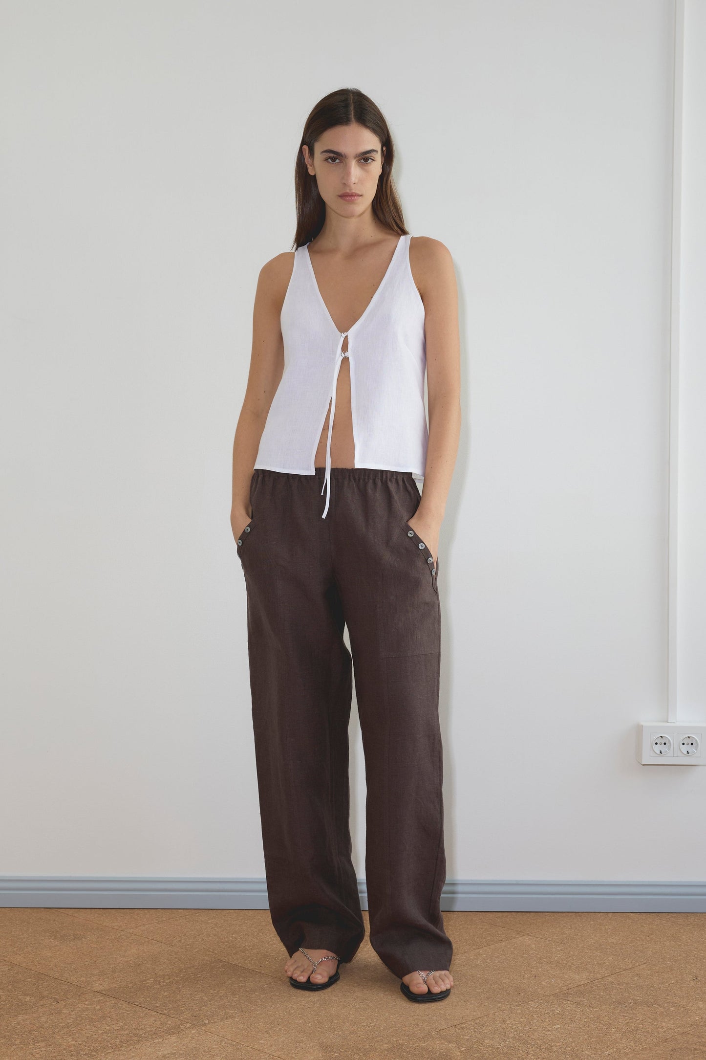 A woman stands indoors against a plain white wall, wearing the Deiji Studios Two Part Top - White with a v-neckline and tie front, paired with loose dark brown pants and sandals. Her hands are in her pockets and her expression is neutral.