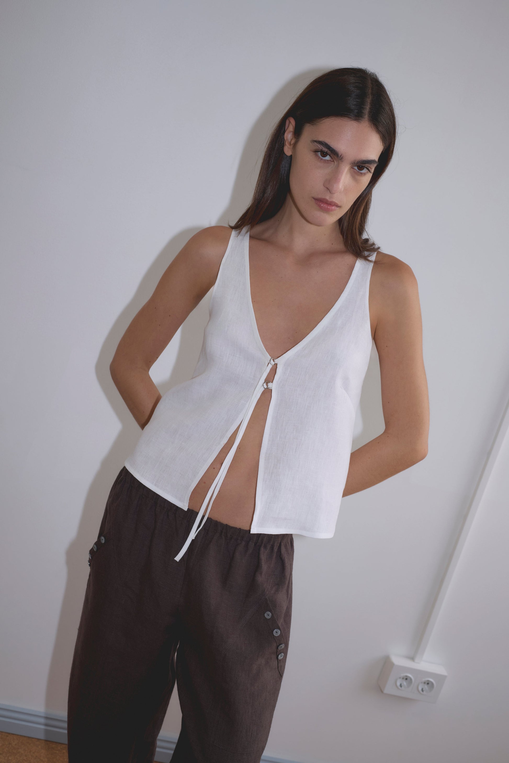 A woman with long, straight brown hair stands against a white wall, wearing Deiji Studios' Two Part Top - White with a v-neckline and front tie, paired with loose dark brown pants. She gazes at the camera with a neutral expression.
