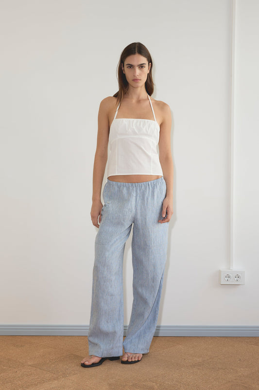 A woman with straight brown hair stands indoors against a white wall, wearing a white halter top and the Angle Pocket Pant - Blue Stripe with black sandals. She has a neutral expression, and a power outlet appears on the wall to her right.