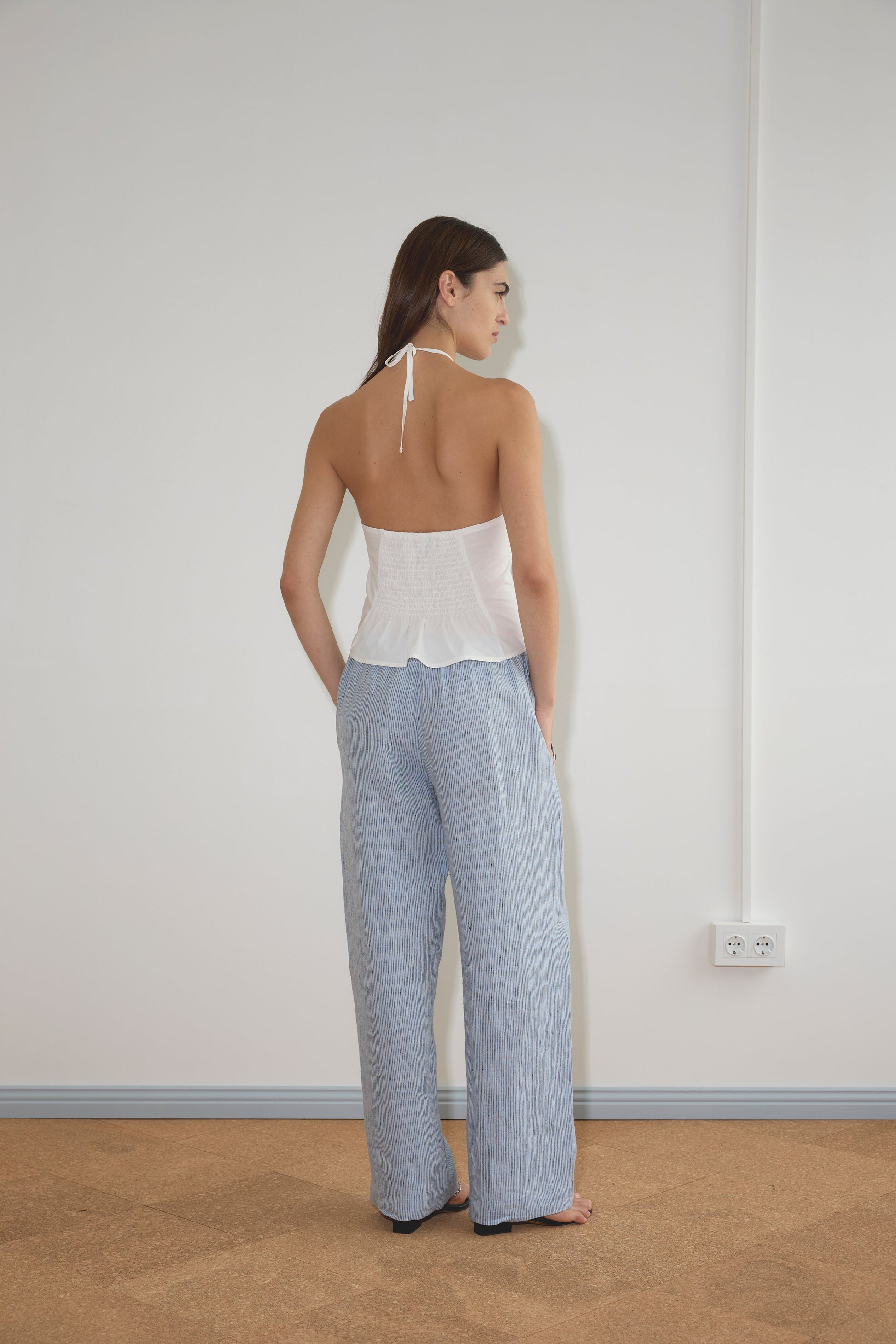 A woman stands facing a white wall, wearing a white halter top and the Angle Pocket Pant - Blue Stripe. Her right hand rests on her hip; a power outlet is visible on the wall.