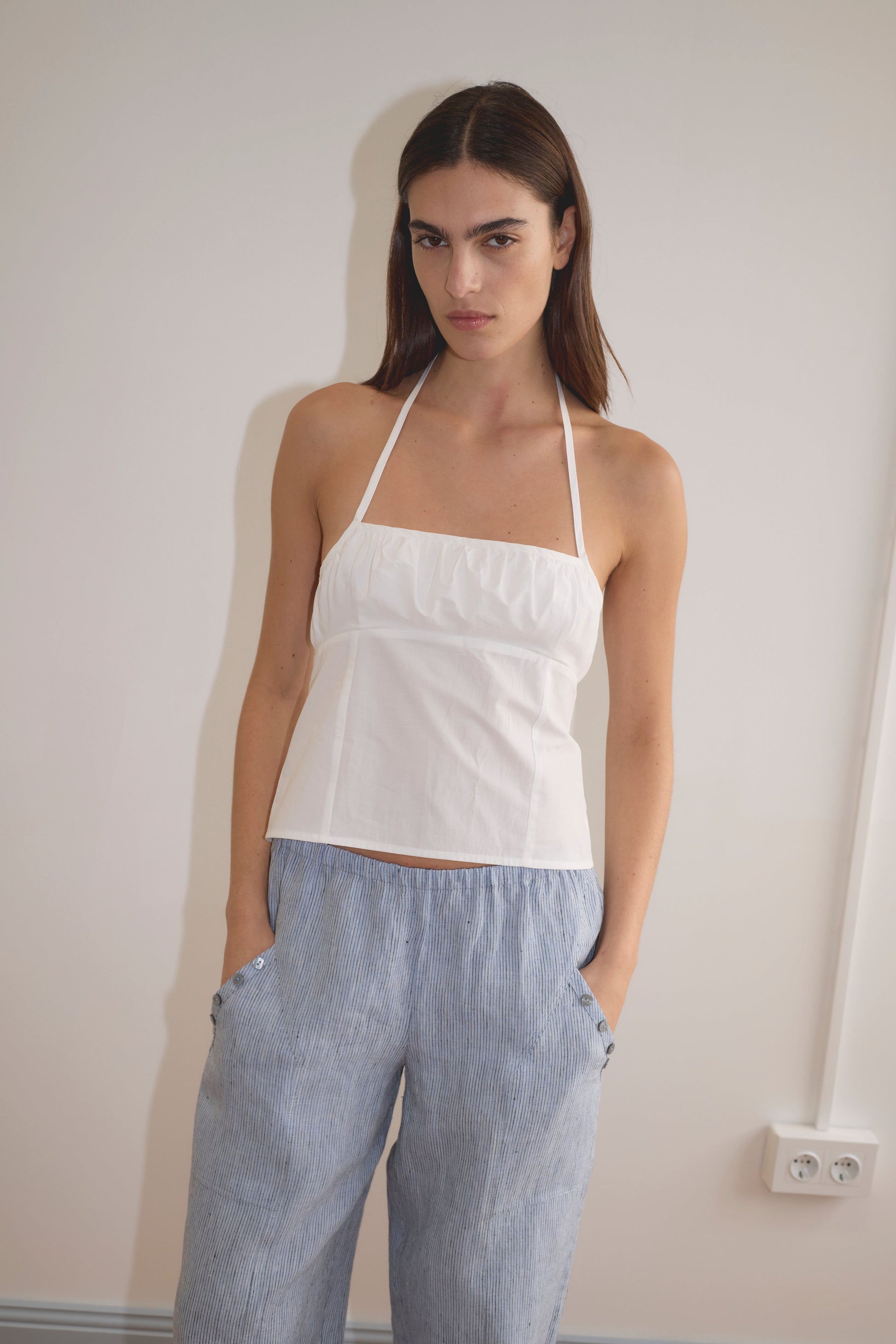 A person with long brown hair stands against a light wall, wearing a white halter top and the Angle Pocket Pant in Blue Stripe, hands in pockets, looking at the camera with a neutral expression.
