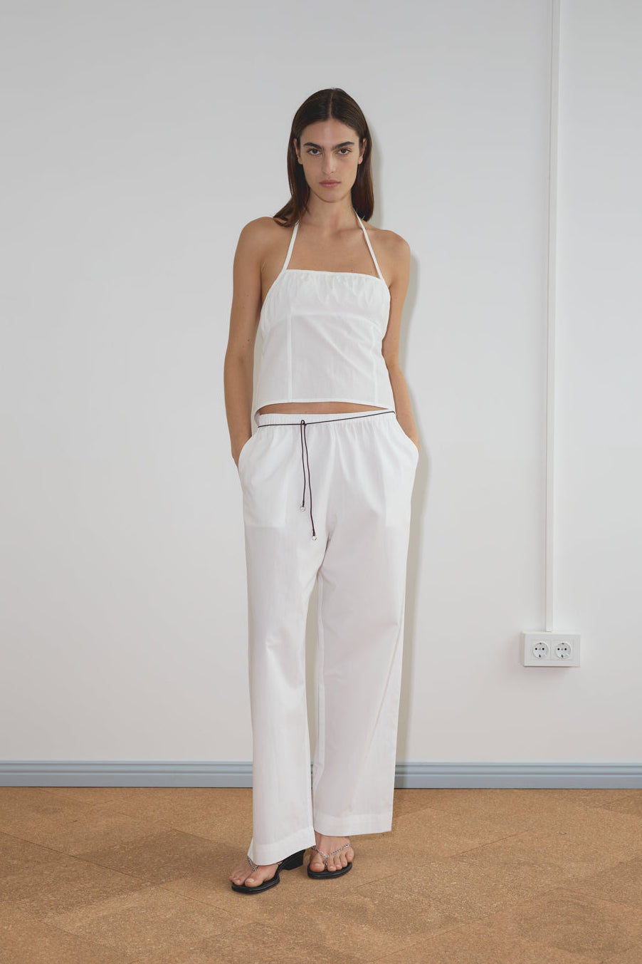 A woman stands indoors against a plain white wall, wearing a white halter-neck camisole, loose recycled polyester pants with side pockets, black sandals, and a stylish Multi Ring Belt in burgundy. Her hands are in her pockets as she looks at the camera.