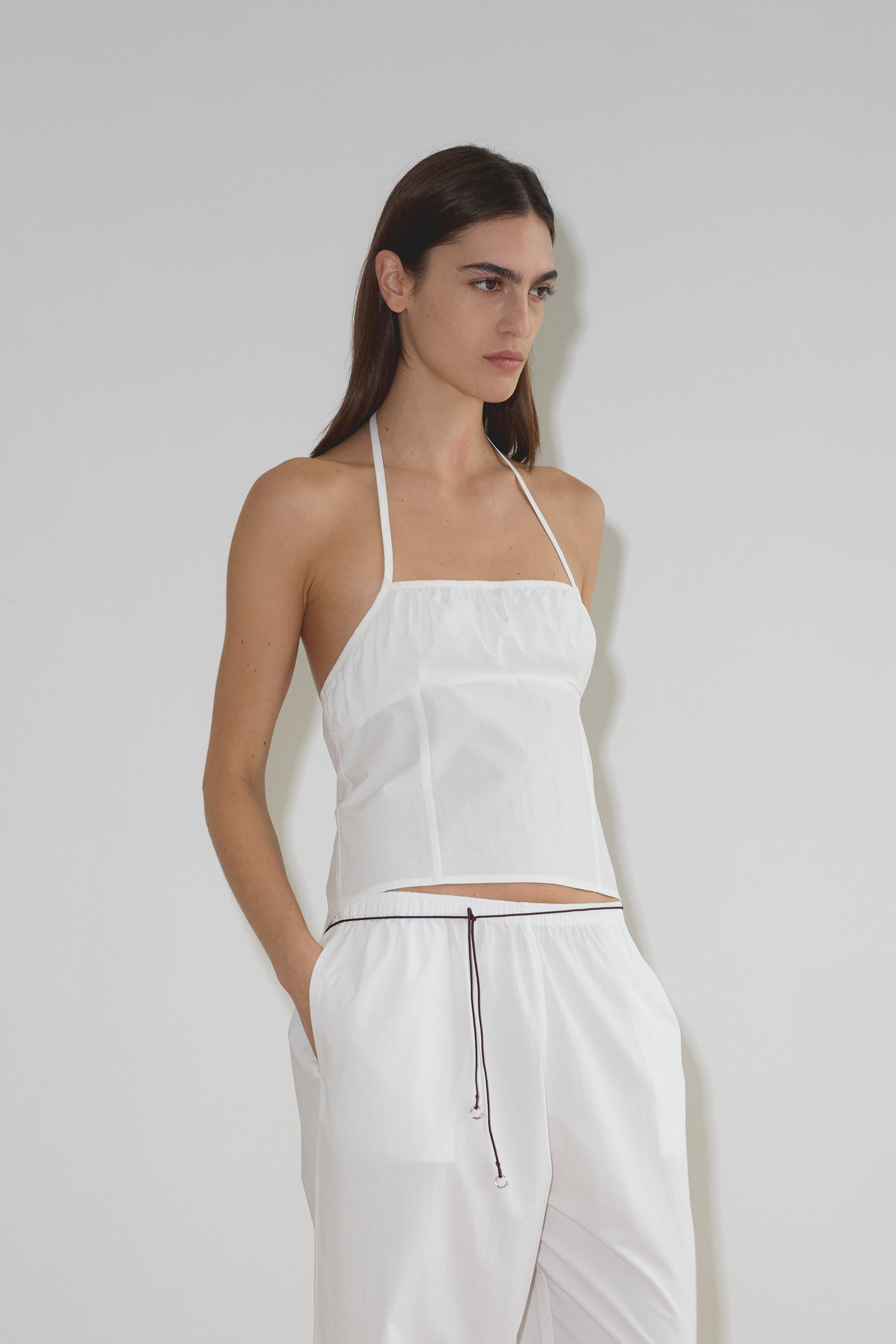 A woman with long dark hair stands against a plain light background, wearing a white halter top and loose white pants styled with a black Multi Ring Belt, her hand in her pocket as she faces slightly to the side.