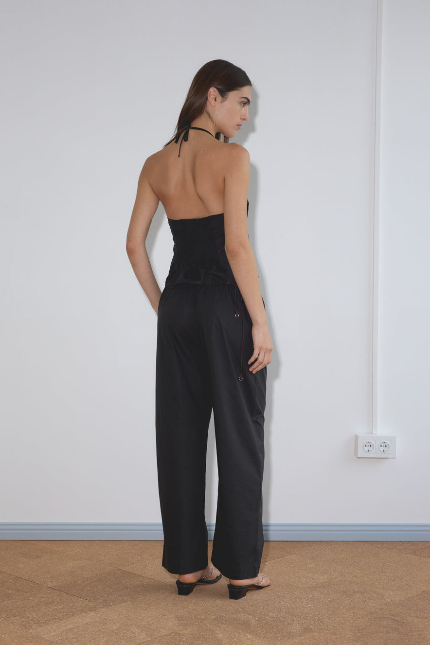 A woman stands indoors facing away, wearing a black halter top, loose black pants with a Multi Ring Belt in burgundy, and black open-toe heels. The white wall behind her features an electrical outlet and blue floor trim.