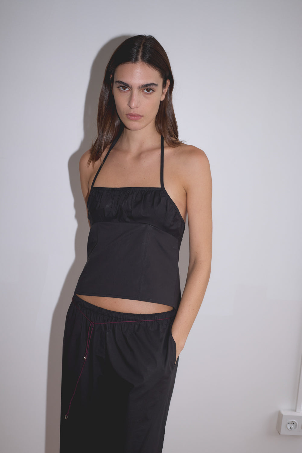 A woman with long dark hair wears a black halter top and pants, styled with the Multi Ring Belt - Burgundy, standing against a white wall with her hand in her pocket and looking directly at the camera.