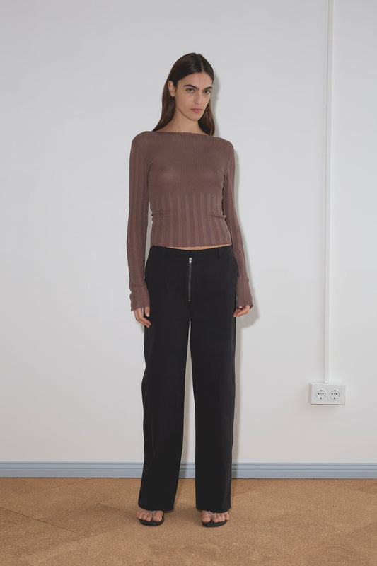 A woman stands indoors against a white wall, wearing a relaxed fit brown top, the Zip Pant - Black, and black open-toe shoes. A double electrical socket and visible cables are on the wall to her right.