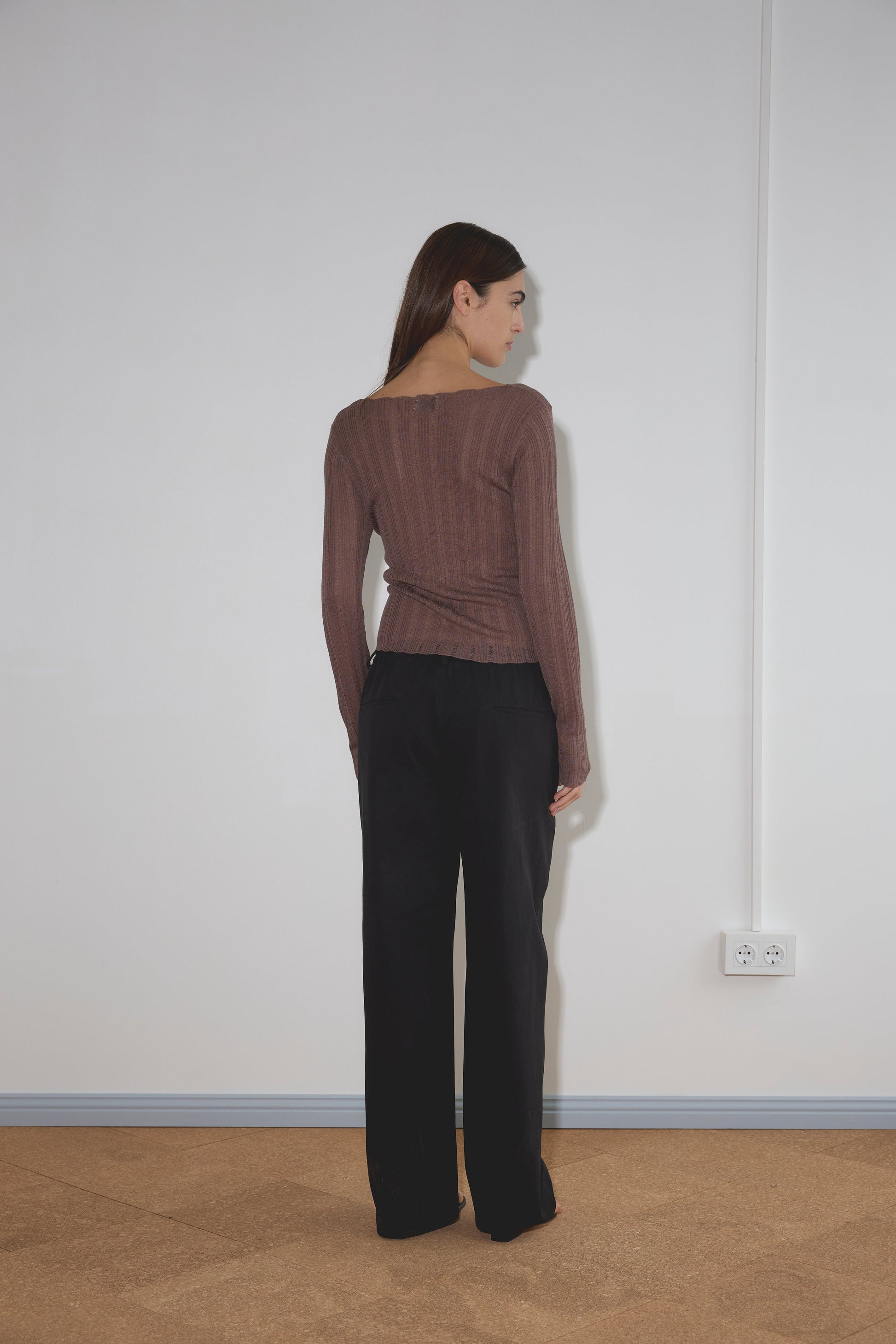 A woman with long brown hair stands facing a white wall, wearing a brown long-sleeve top and the Zip Pant - Black by Deiji Studios. A power outlet is visible on the wall near her right side.