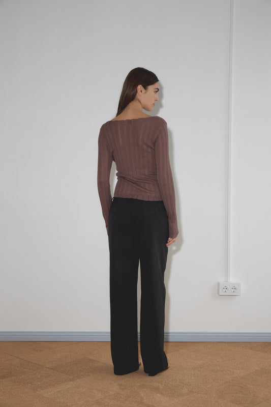 A woman with long brown hair stands facing a white wall, wearing a brown long-sleeve top and the Zip Pant - Black by Deiji Studios. A power outlet is visible on the wall near her right side.