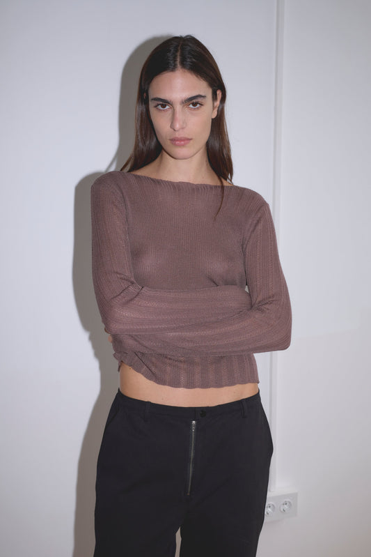 A person with long dark hair stands against a white wall, wearing a sheer brown long-sleeve top and the Zip Pant - Black in organic cotton twill, arms crossed, looking directly at the camera with a neutral expression.