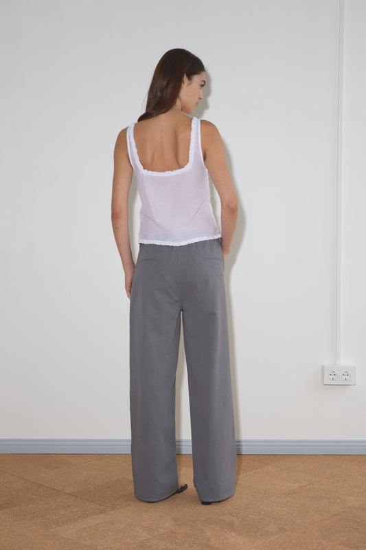 A woman with straight brown hair faces a white wall, wearing the Button Frill Top - White from Deiji Studios and loose gray pants. Light brown flooring and electrical outlets are visible on the wall to her right.