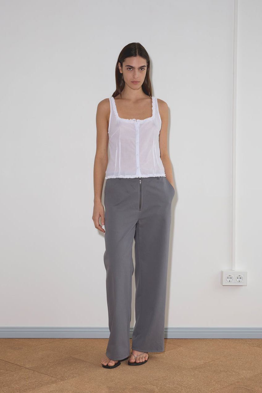 A woman stands indoors against a white wall, wearing the Button Frill Top - White by Deiji Studios with lace trim, loose gray trousers, black sandals, and her hands in her pockets. She has straight dark hair.