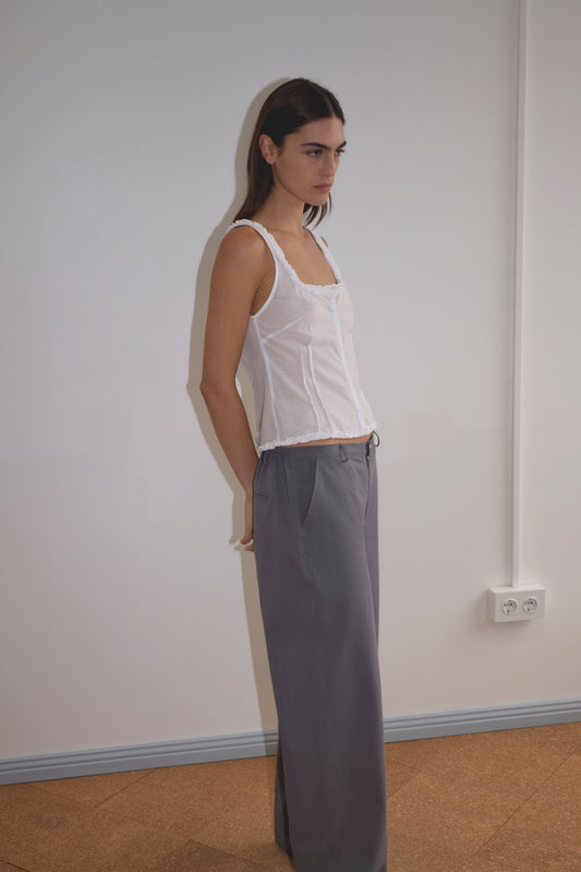A woman with long brown hair stands indoors against a white wall, wearing the Deiji Studios Button Frill Top in white and loose gray trousers. She gazes slightly to the side, her arms relaxed behind her back.