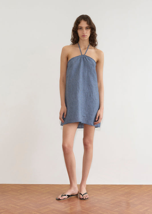 A person with wavy brown hair wears the Cross Over Dress, a short strapless blue and white striped design, paired with black sandals, standing indoors on a wooden floor against a plain white wall.