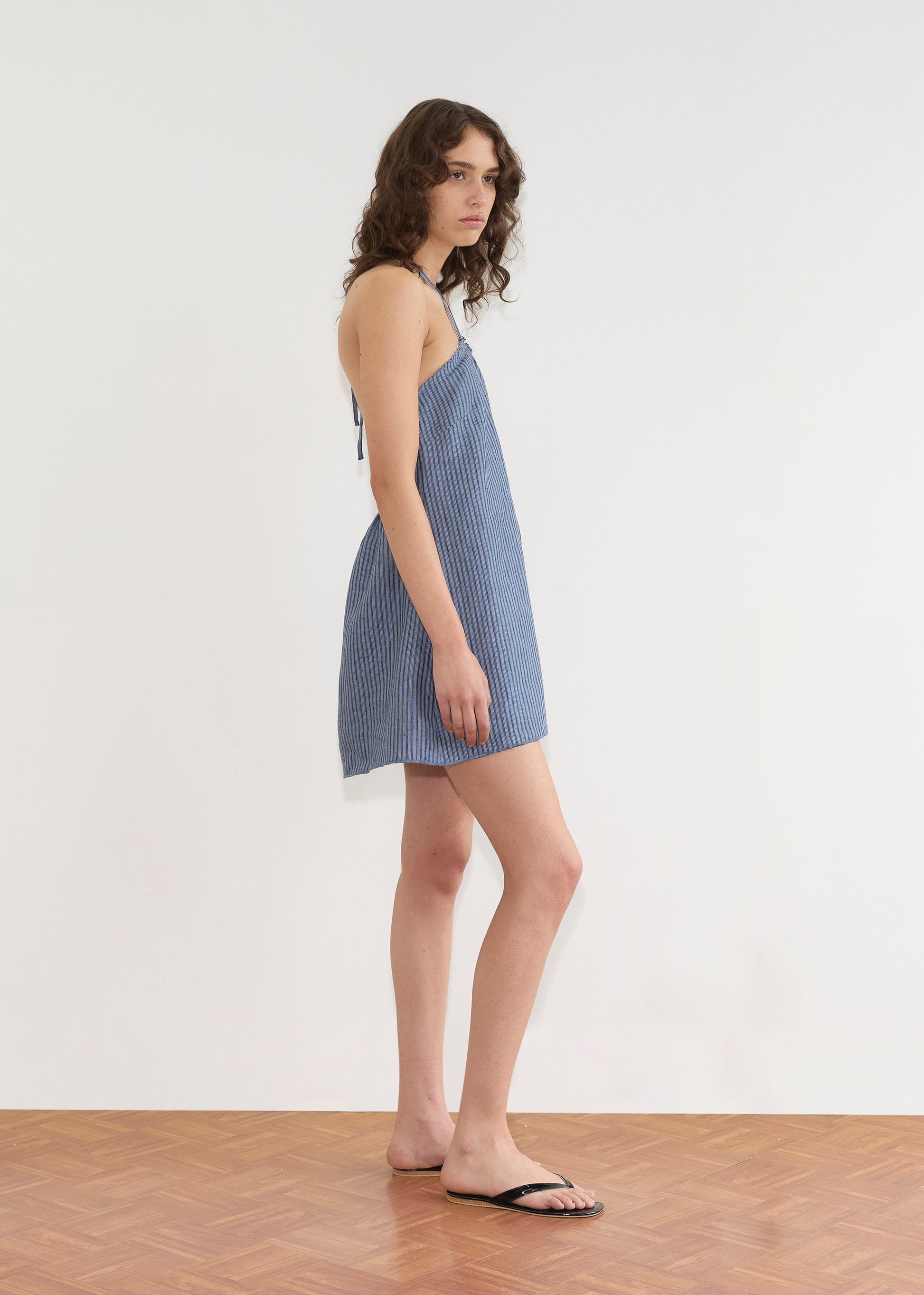 A person with wavy brown hair wears the Deiji Studios Cross Over Dress - Azure Stripe and black sandals, standing on a wooden floor against a plain white wall, facing slightly to the side.