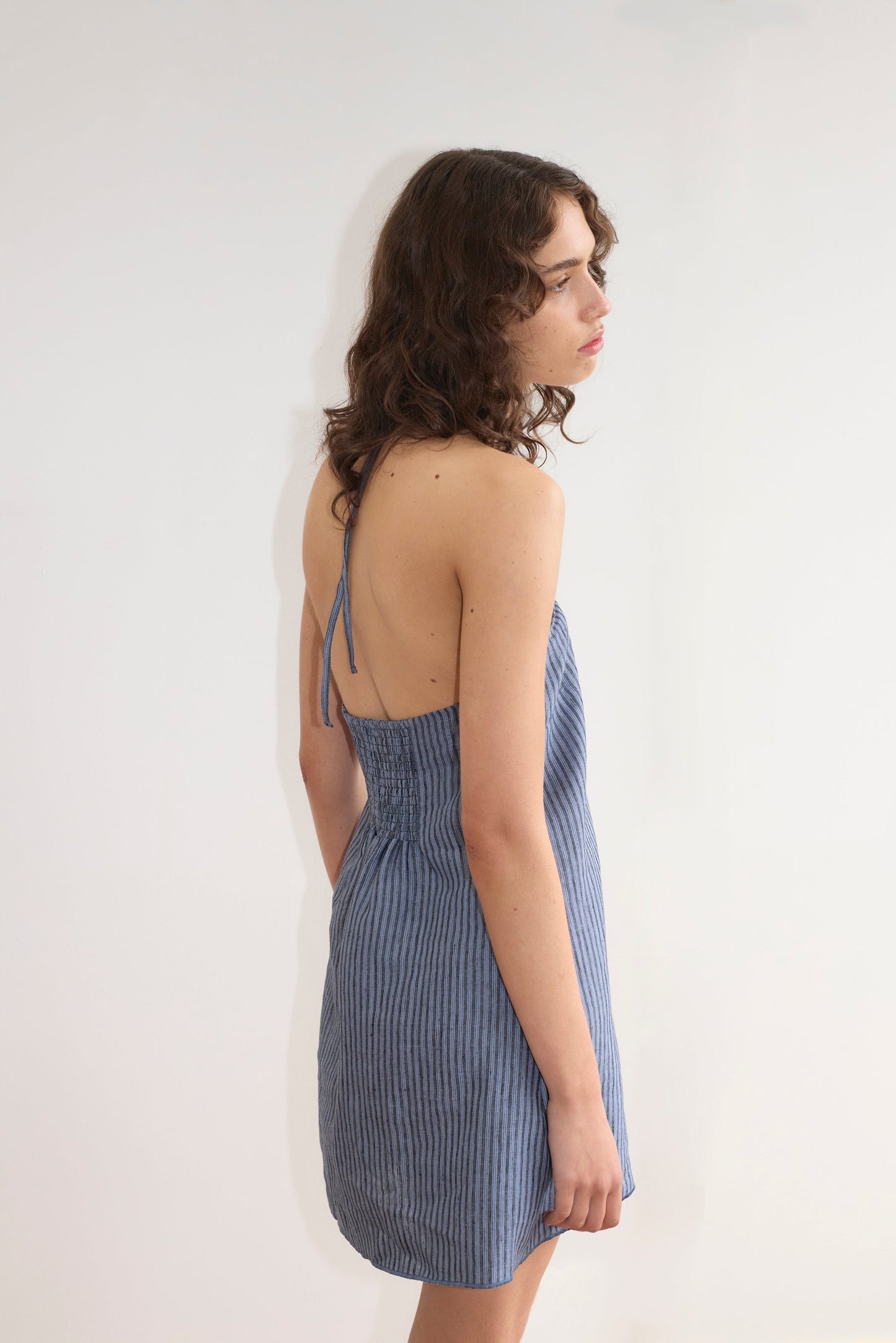 A woman with wavy brown hair models the Cross Over Dress - Azure Stripe from Deiji Studios. She stands sideways in the blue, backless, striped dress and gazes to the right against a plain white background.