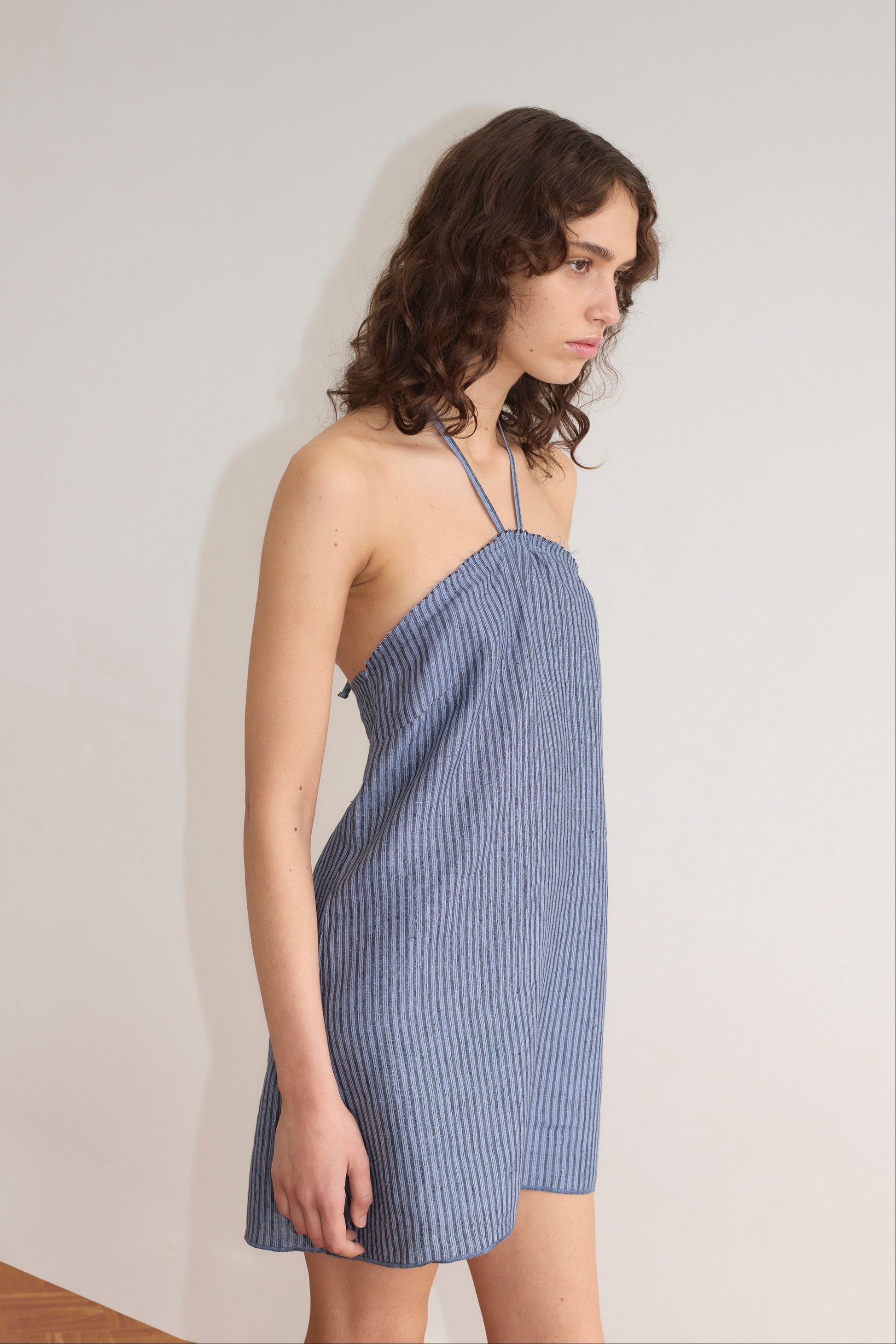 Against a plain white wall, a person with wavy brown hair wears Deiji Studios’ Cross Over Dress - Azure Stripe—a sleeveless, relaxed mini dress featuring a halter neck and subtle vertical stripes.