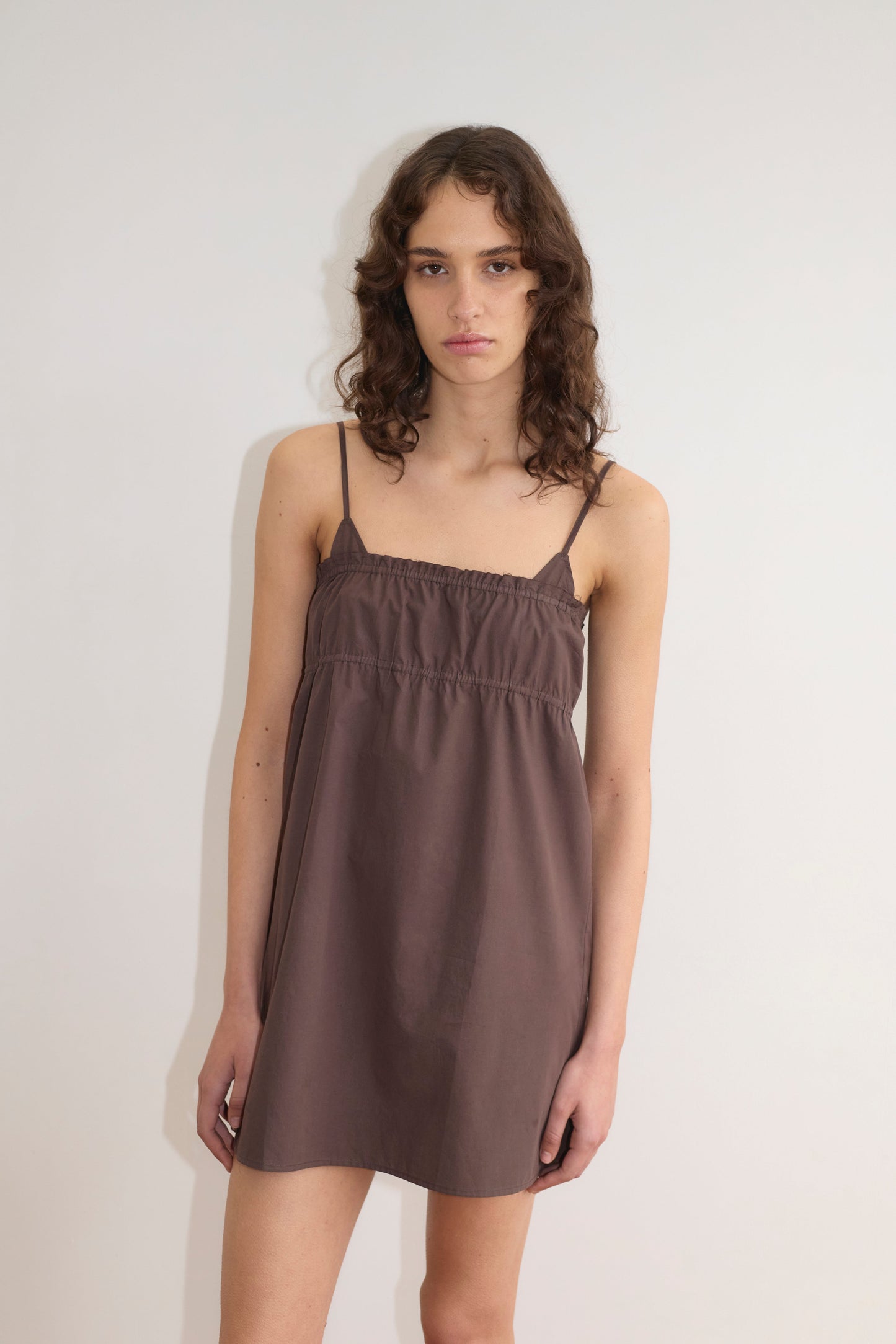 A person with long, wavy brown hair wears the Edge Dress - Sepia from Deiji Studios—a sleeveless, lightweight dress with thin straps—standing against a white wall and looking at the camera with a neutral expression.