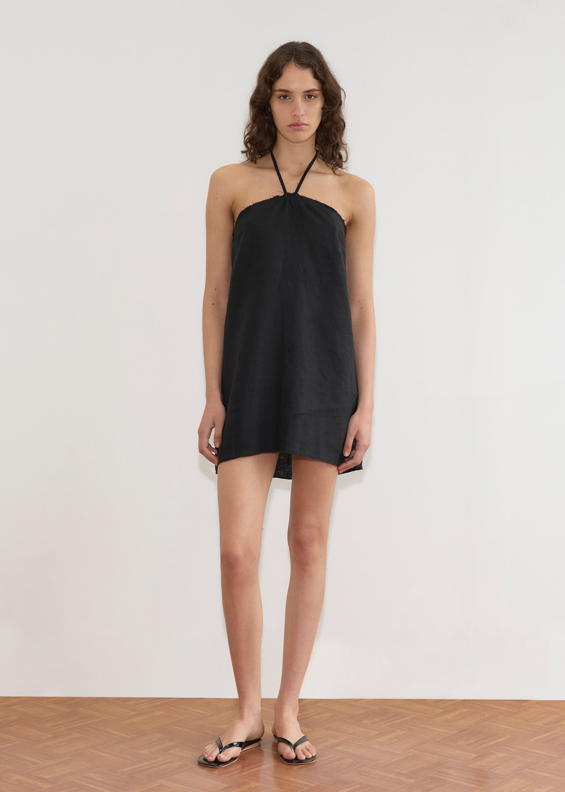 A person with wavy brown hair stands on a wood floor against a plain white wall, facing forward with a neutral expression. They wear the Cross Over Dress - Black from Deiji Studios and black sandals.