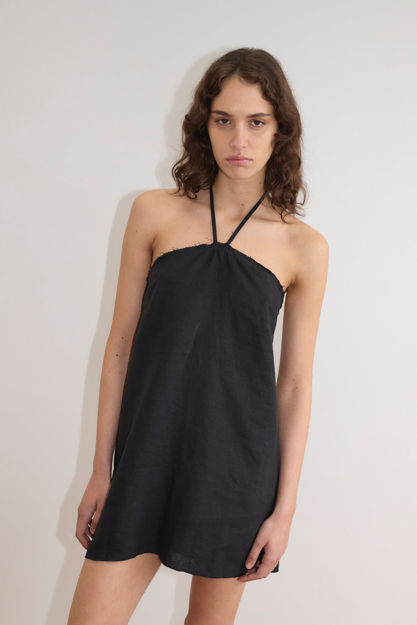 A person with long, wavy brown hair wears the Cross Over Dress - Black by Deiji Studios and stands against a plain white wall, looking at the camera with a neutral expression.