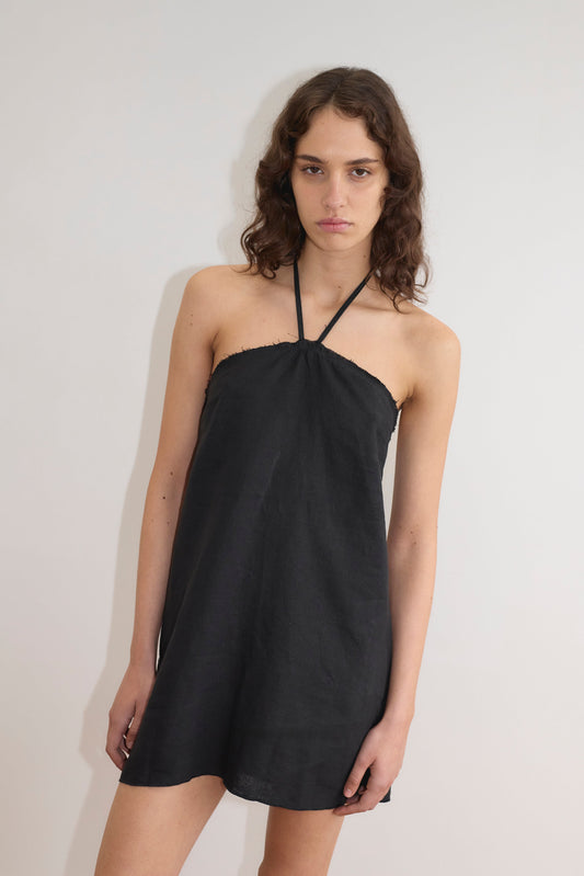 A person with long, wavy brown hair wears the Cross Over Dress - Black by Deiji Studios and stands against a plain white wall, looking at the camera with a neutral expression.